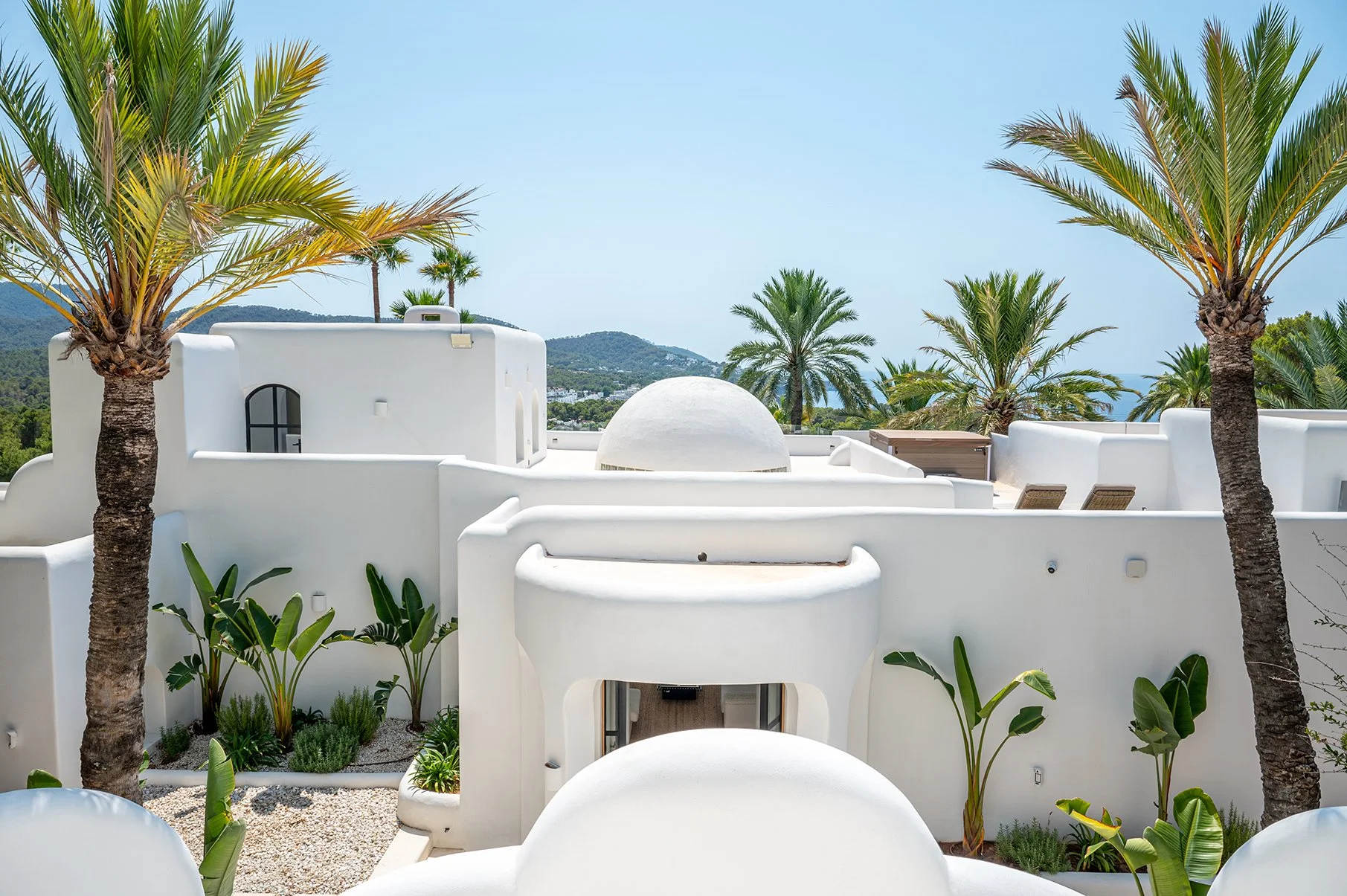 White Mediterranean-style building with rounded domes and walls, surrounded by palm trees and lush plants, under a clear blue sky.