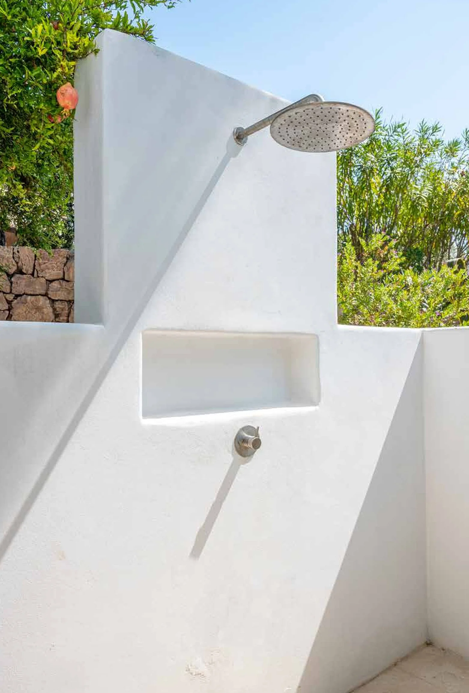 Outdoor shower with a large rain showerhead, a soap or shampoo niche, and a handheld shower attached to a white wall, with greenery and a blue sky in the background.