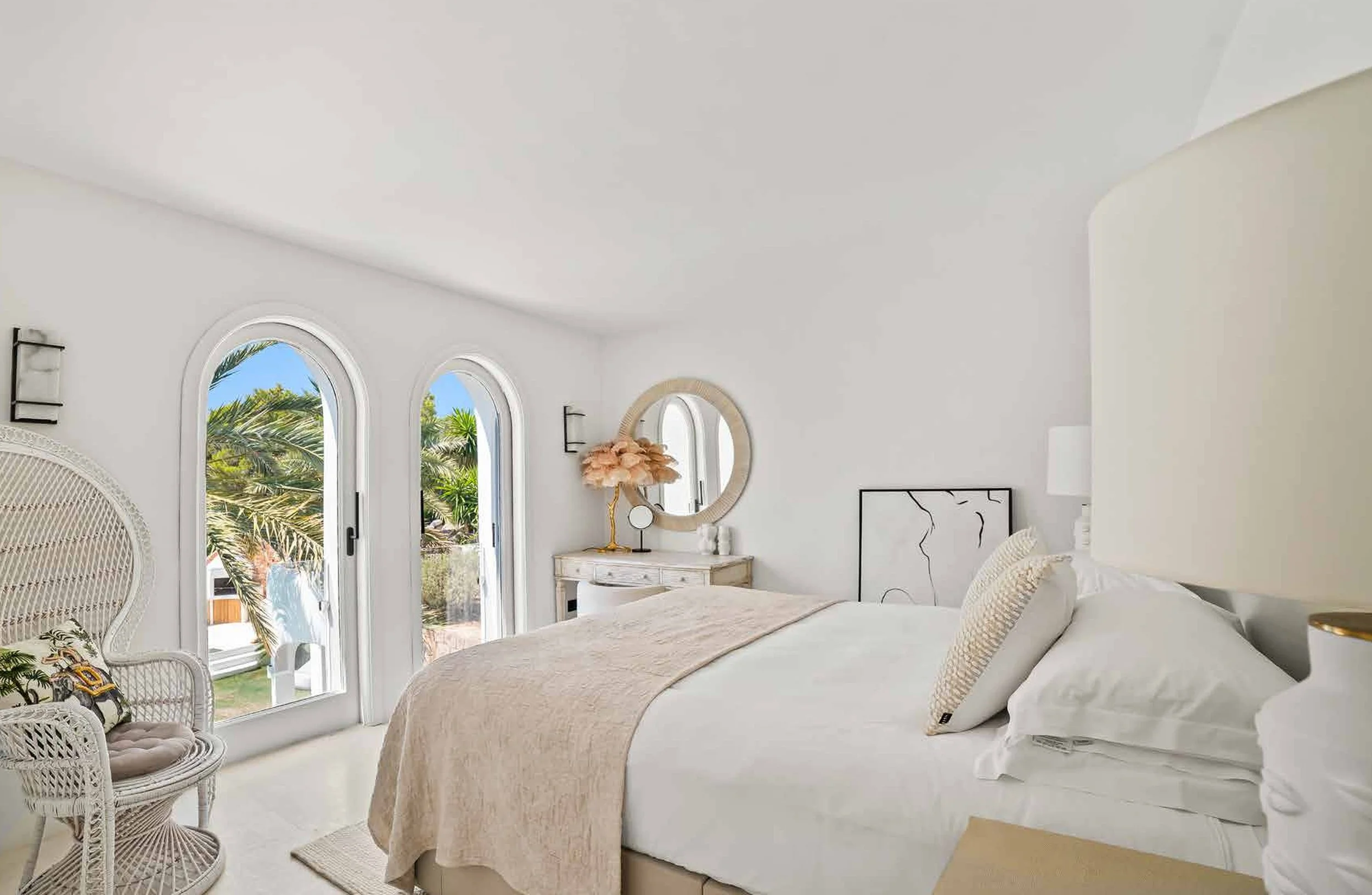 Bright bedroom with white walls, arched windows showing palm trees outdoors, beige bedding, decorative lamp with pink flowers, round mirror, and modern art leaning against the wall.