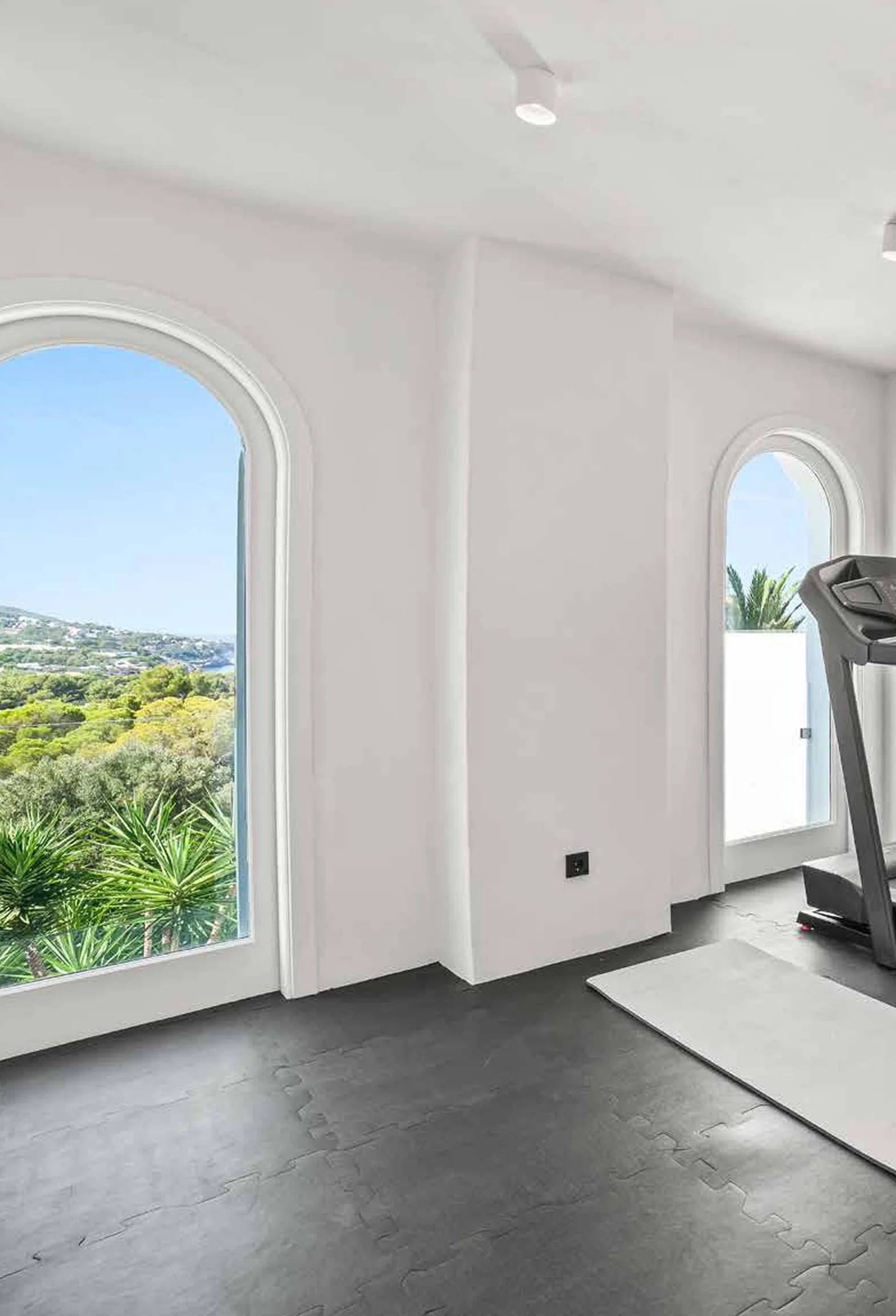 A minimalistic home gym with two large arched windows showing a green landscape outside, a treadmill near the right window, a white mat on the floor, and white walls.