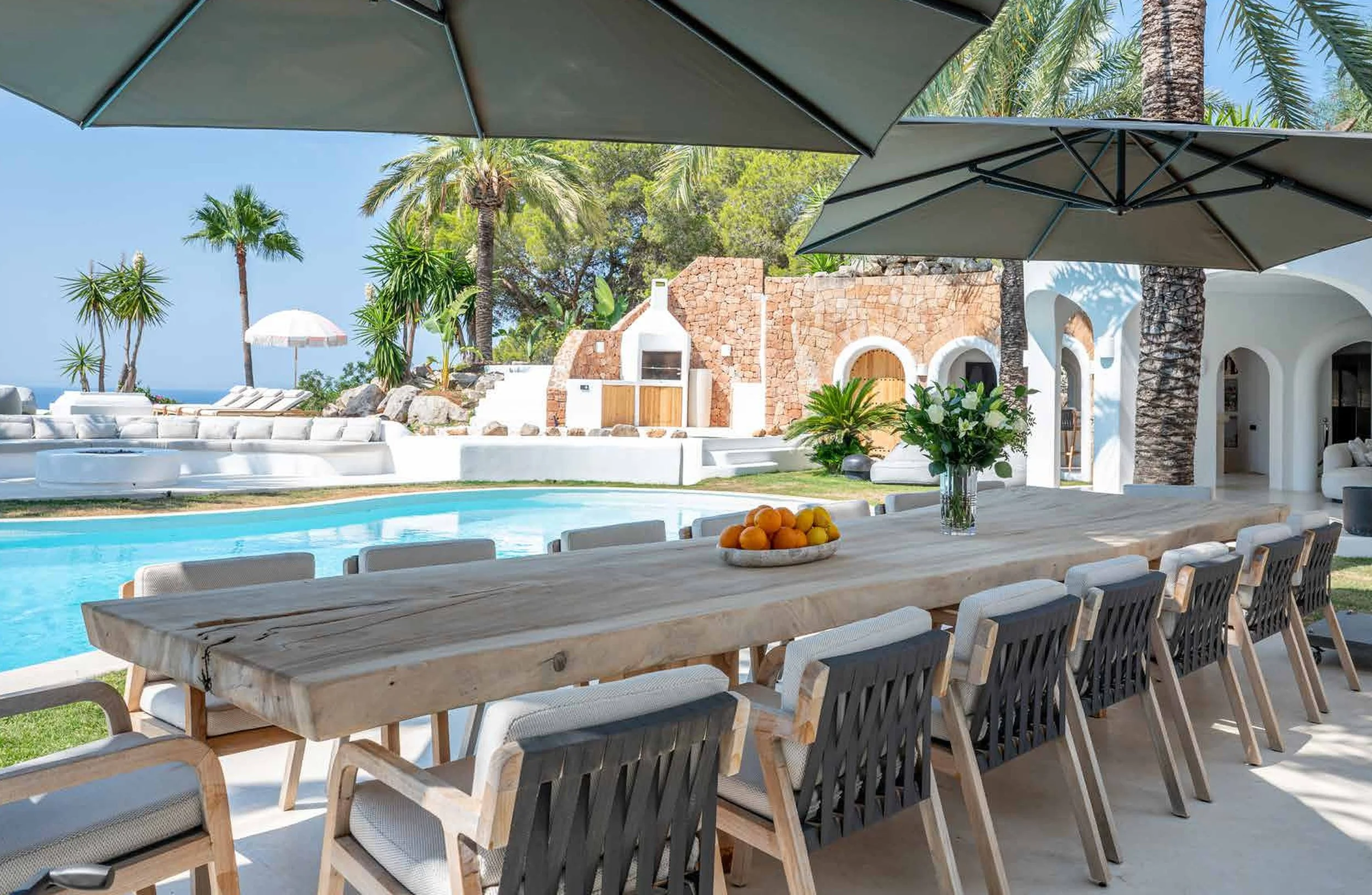 Outdoor poolside dining area with a long wooden table, white chairs, large umbrellas, a glass vase with flowers, a bowl of oranges, and a view of a swimming pool surrounded by palm trees and tropical plants.
