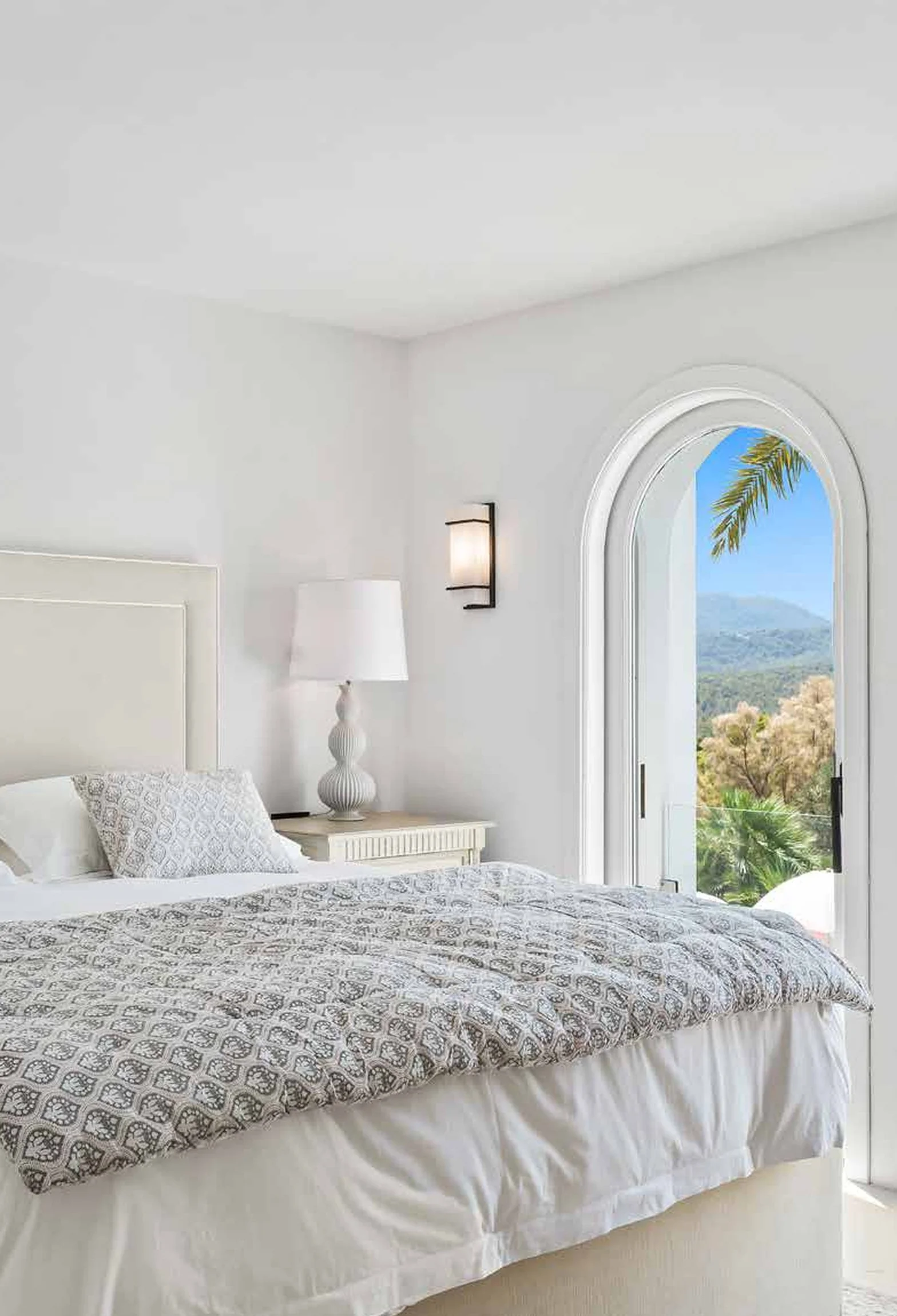 A bright bedroom with white walls, a bed with patterned bedding, a white headboard, a white bedside table with a decorative lamp, a wall-mounted light fixture, and an arched glass door revealing a scenic view of mountains and greenery with a palm tre