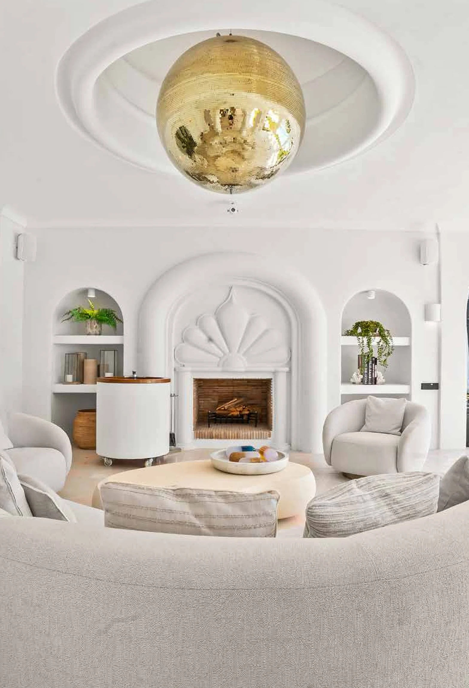 Living room with white walls, a white armchair, a white sofa, a cream-colored coffee table with decorative items, built-in shelves with plants and books, and a white fireplace with a decorative arch. A round, gold reflective ceiling light fixture han