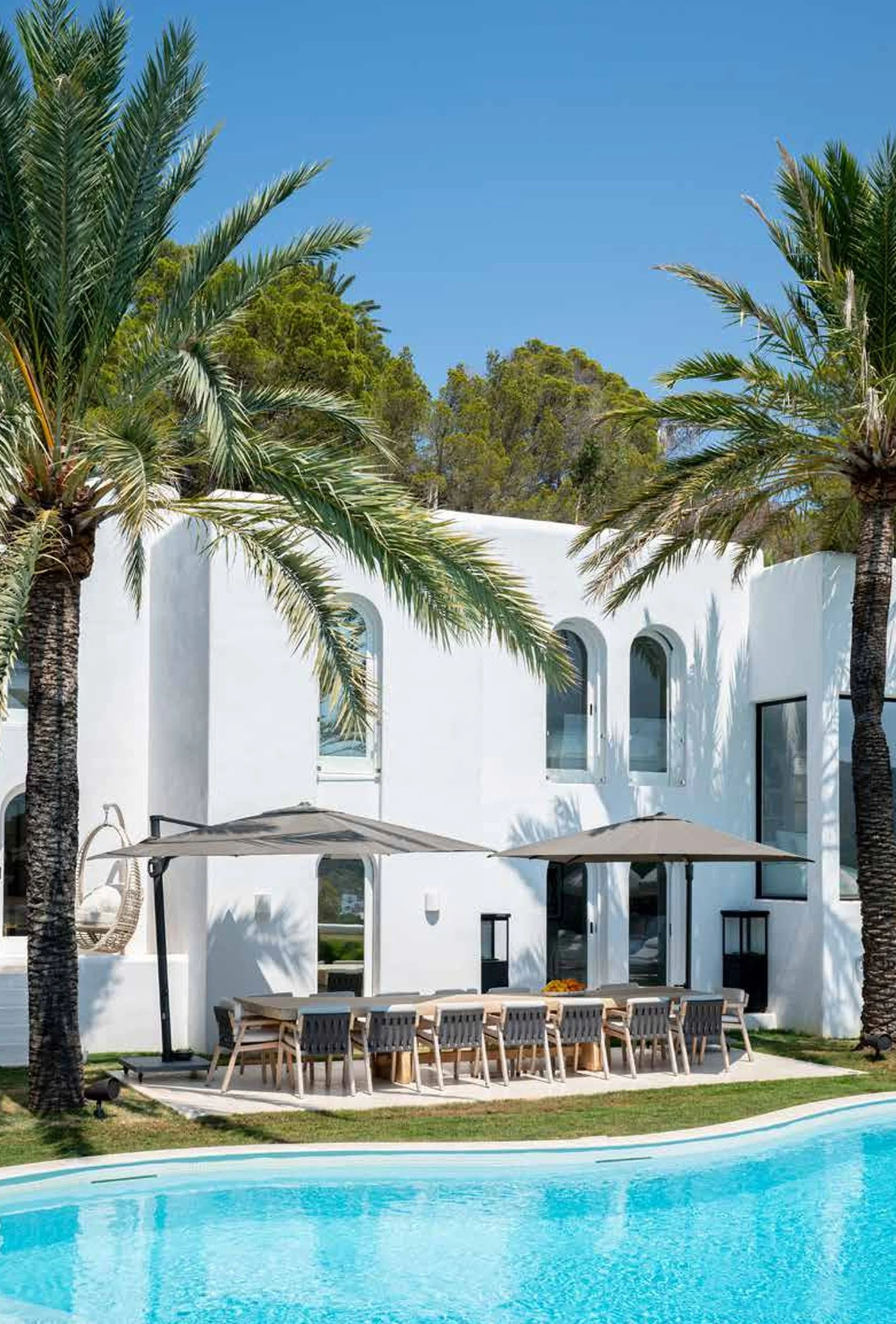 A white modern house with arched windows, surrounded by palm trees, with a swimming pool and outdoor dining area with umbrellas.