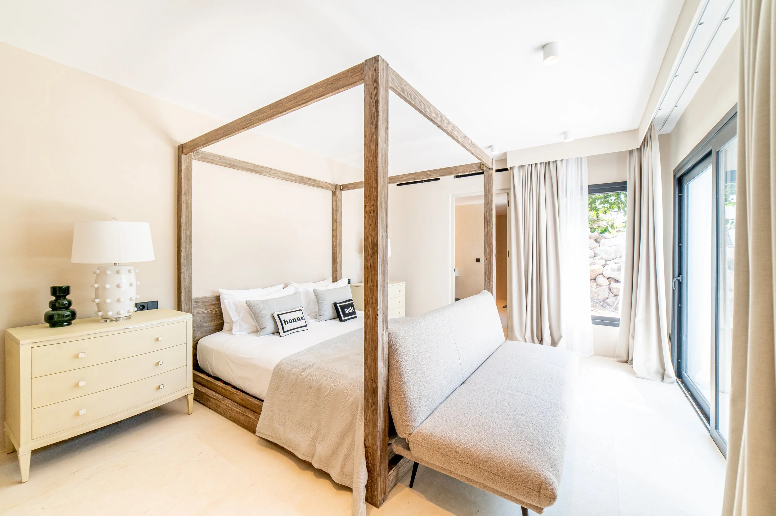 A bedroom with a four-poster bed, dresser, and sliding glass door with curtains, overlooking a rocky landscape outside.