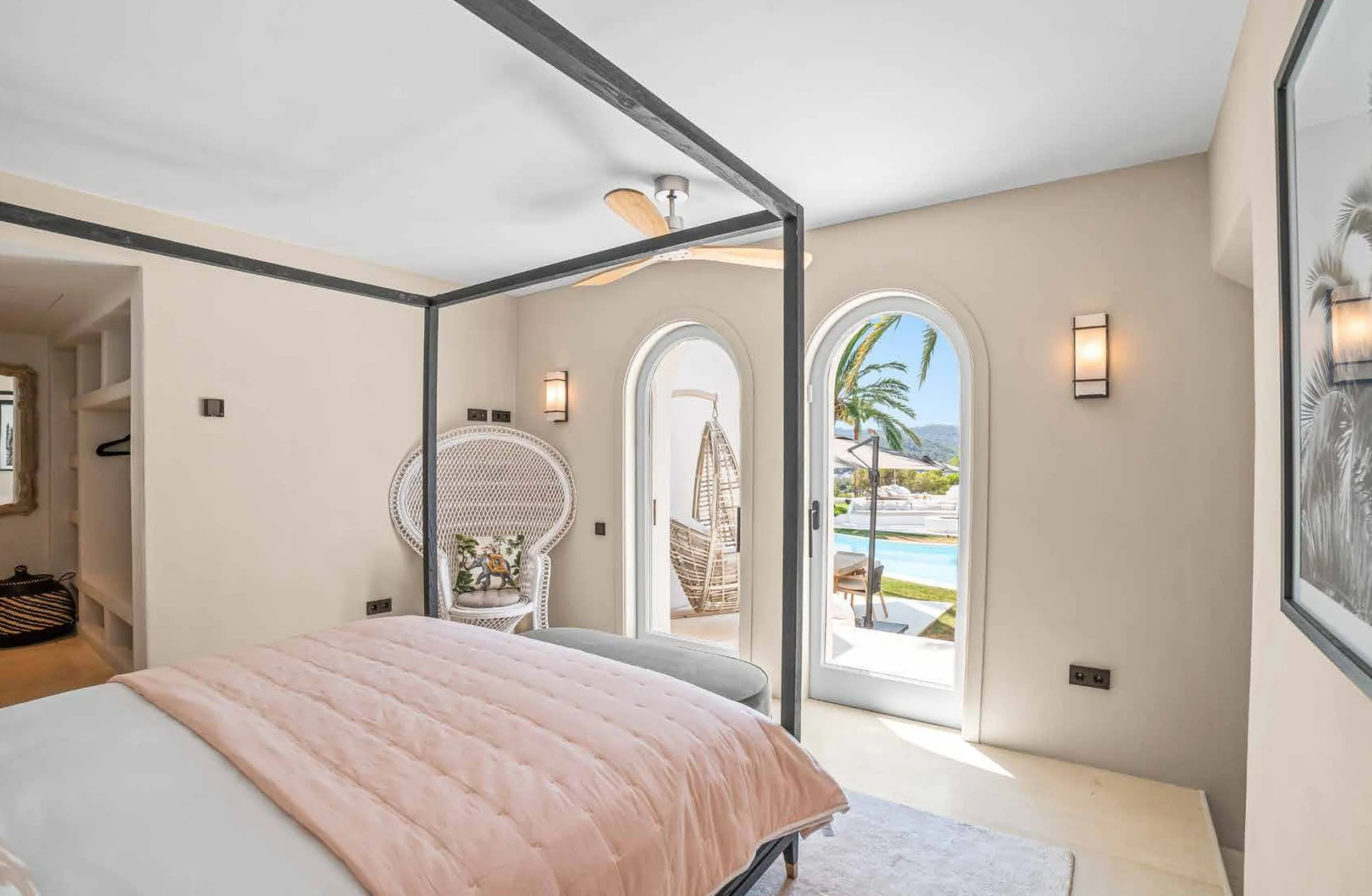 A bedroom with a four-poster bed, pink bedding, a peacock chair with a decorative pillow, and arched glass doors leading to a patio with a pool, lounge chairs, and palm trees beyond.
