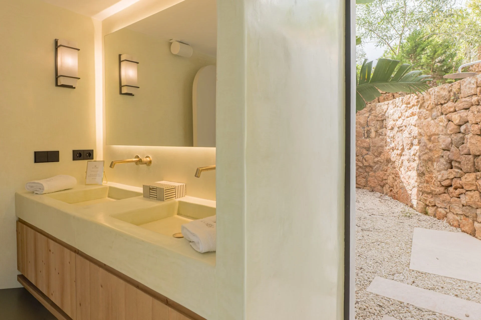 Bathroom with a beige sink, towel, and mirror, separated from an outdoor garden area with stone wall and green plants by a glass wall.