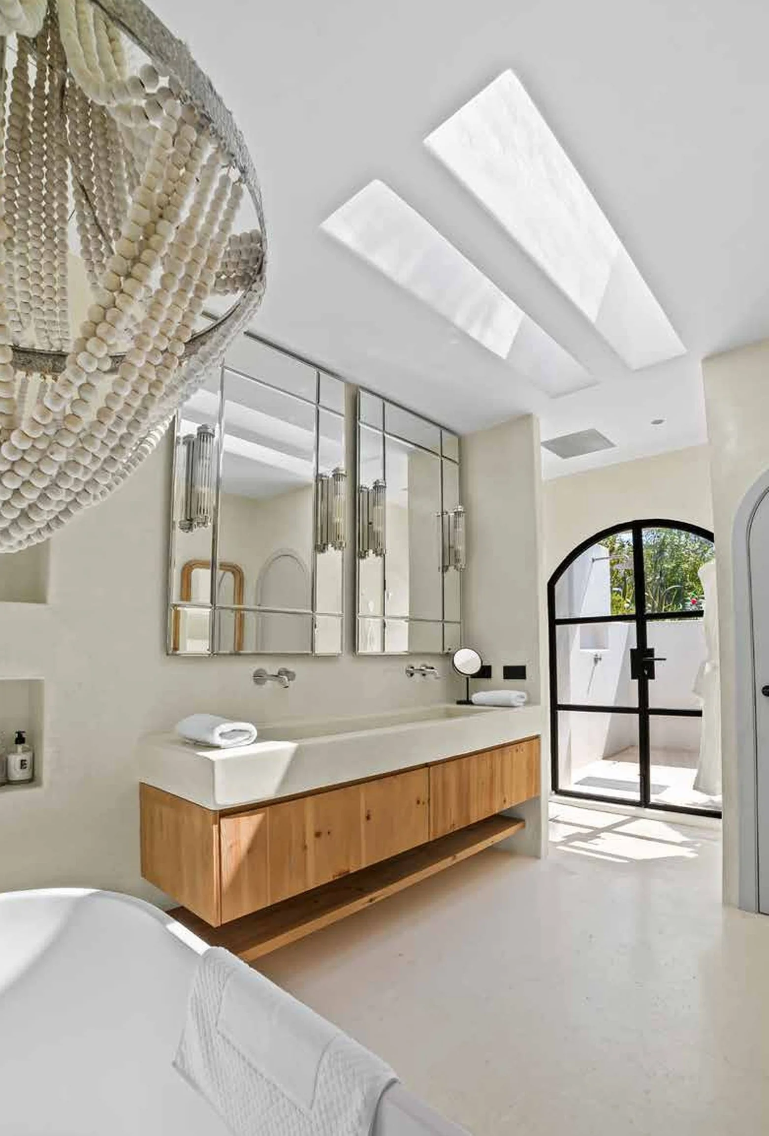 Modern bathroom with cream-colored walls, large mirrors, wooden vanity, hanging towels, black arched door, and outdoor view.