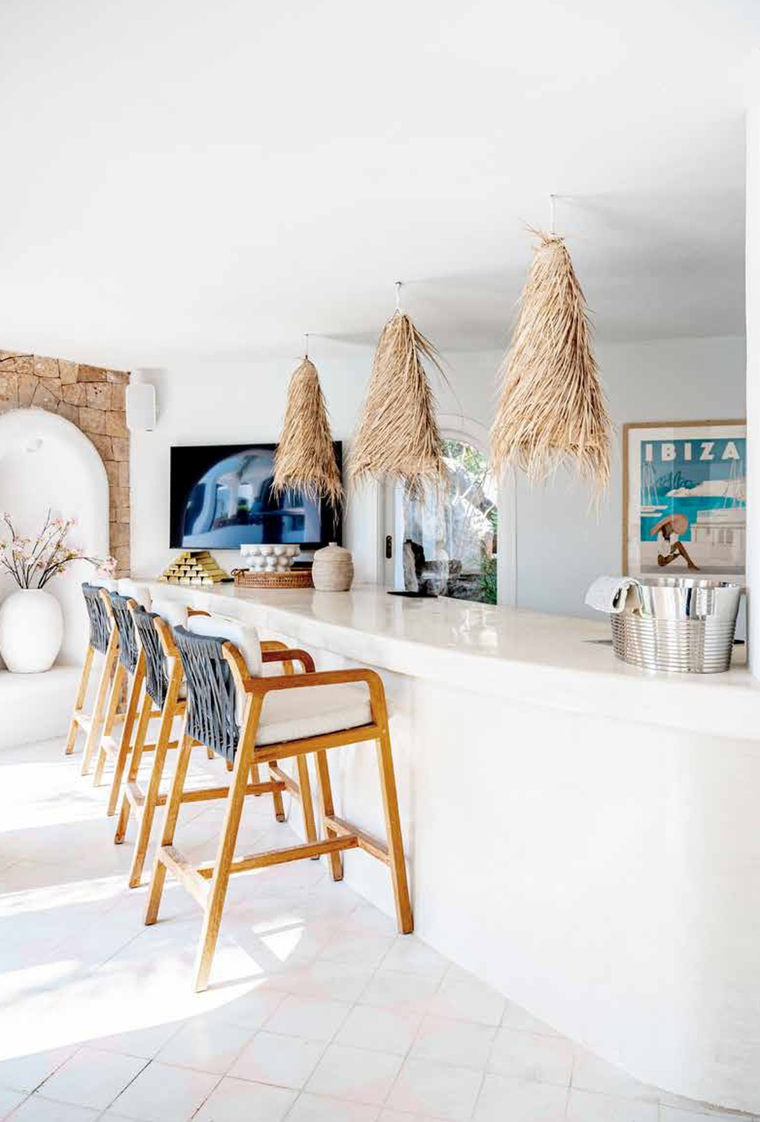 Bright, modern kitchen bar area with four wooden barstools featuring white cushions. The kitchen has a white marble countertop, a flat-screen TV mounted on the wall, and decorative items including dried grass pendant lights, a framed Ibiza poster, an