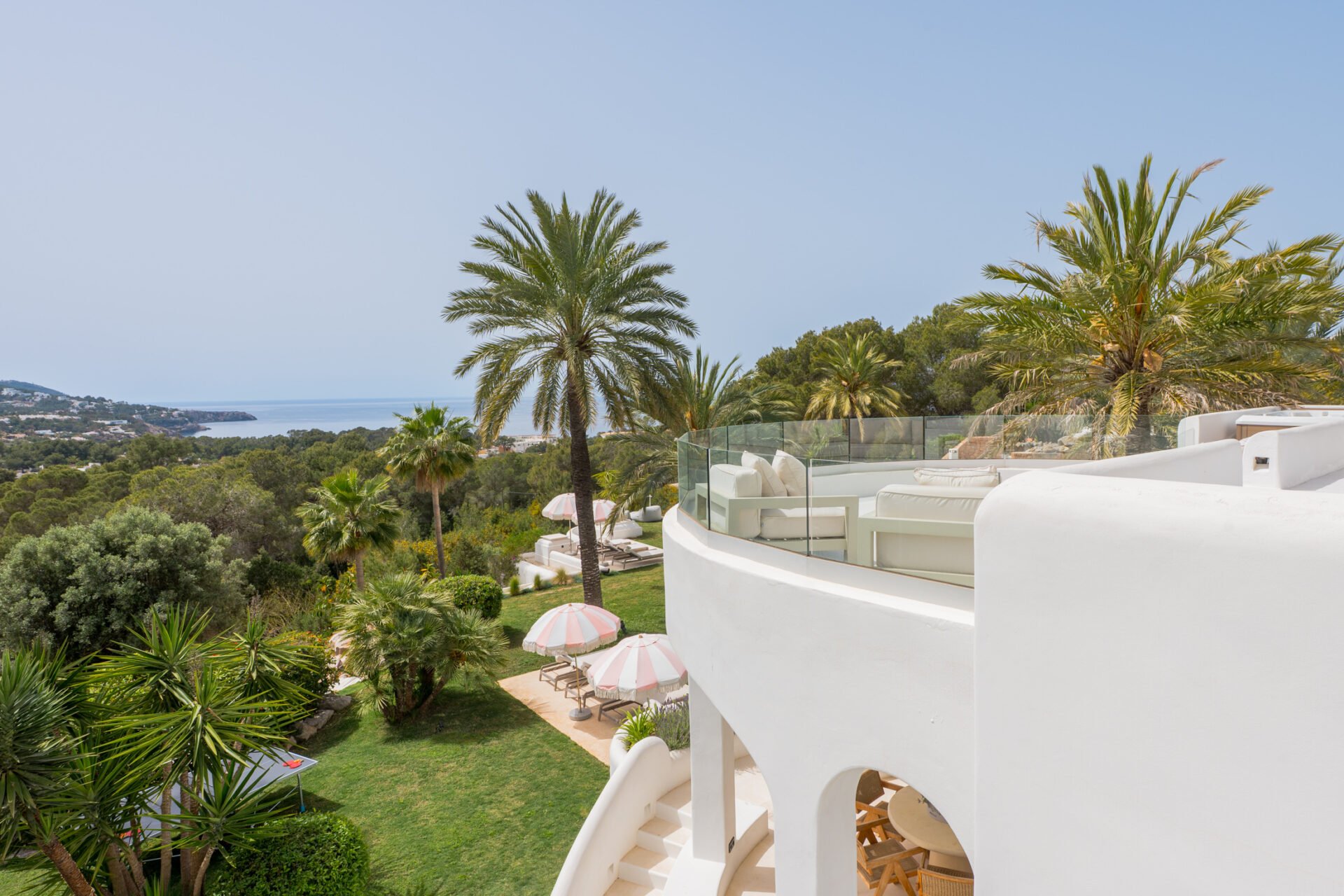 View of a white modern building with outdoor patio and glass railing, surrounded by lush greenery and palm trees, overlooking a distant coastline and ocean under a clear sky.