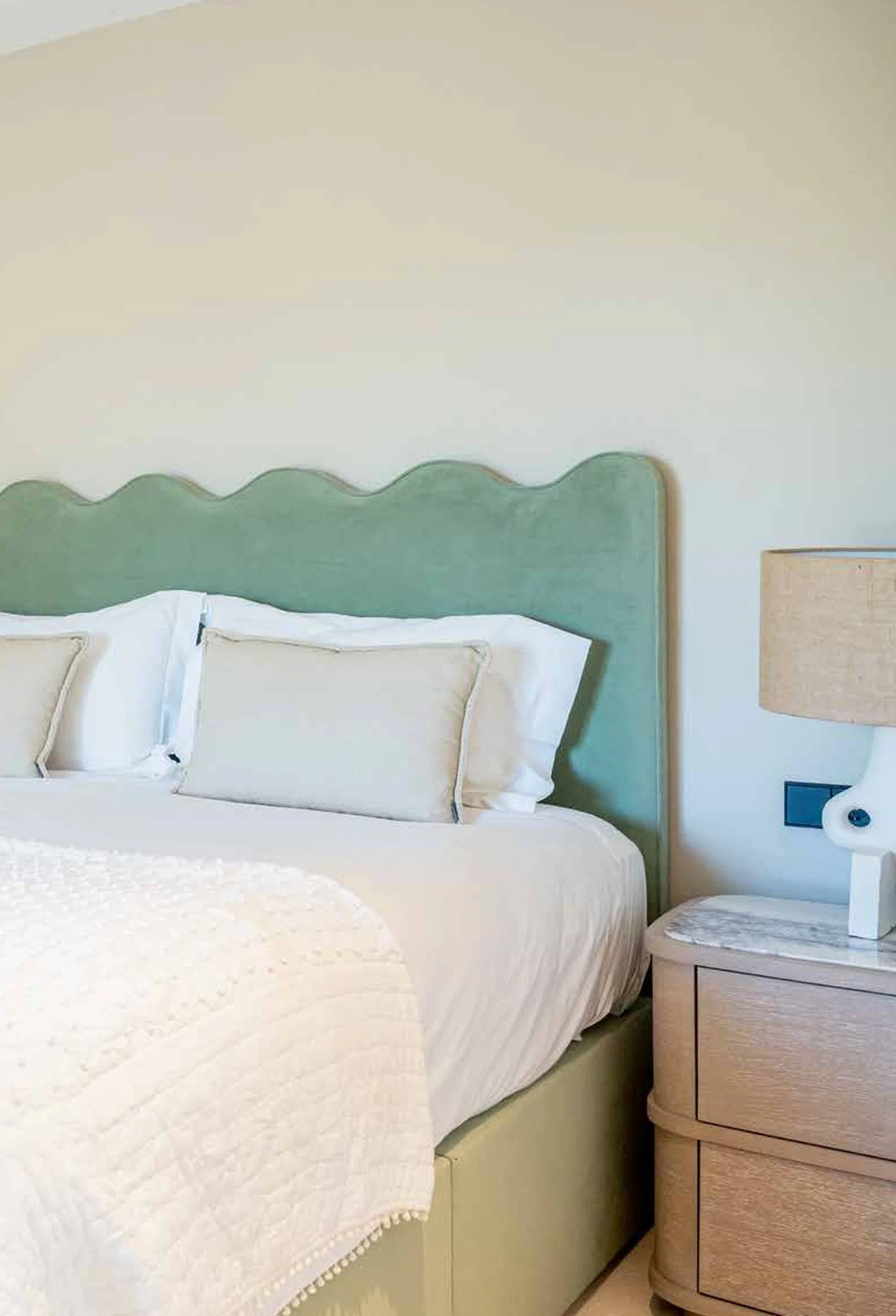 A neatly made bed with white pillows, a light-colored quilt, and a teal upholstered headboard, with a wooden nightstand and a beige lamp beside the bed.