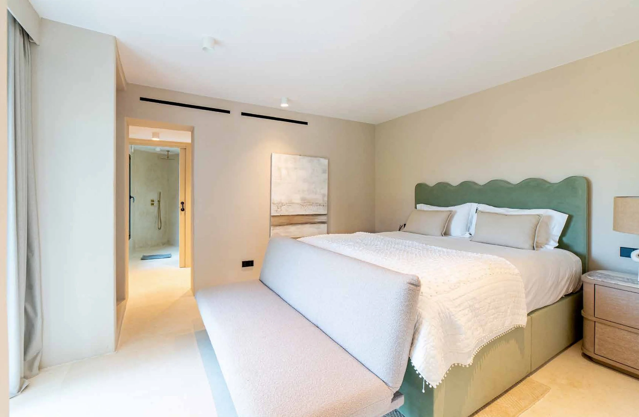 A hotel room with a large bed, green headboard, and white bedding, adjacent to a nightstand with a beige lamp. There is a bench at the foot of the bed and a doorway leading to a bathroom with a visible shower.