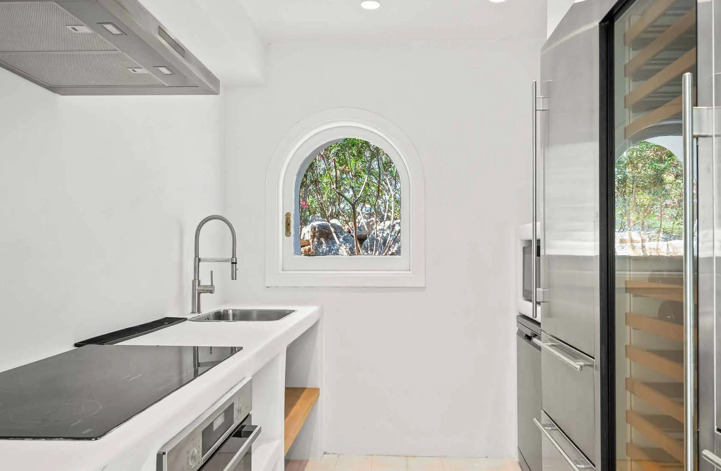Modern kitchen with white walls, stainless steel appliances, a small arched window showing trees outside, and minimalist design.