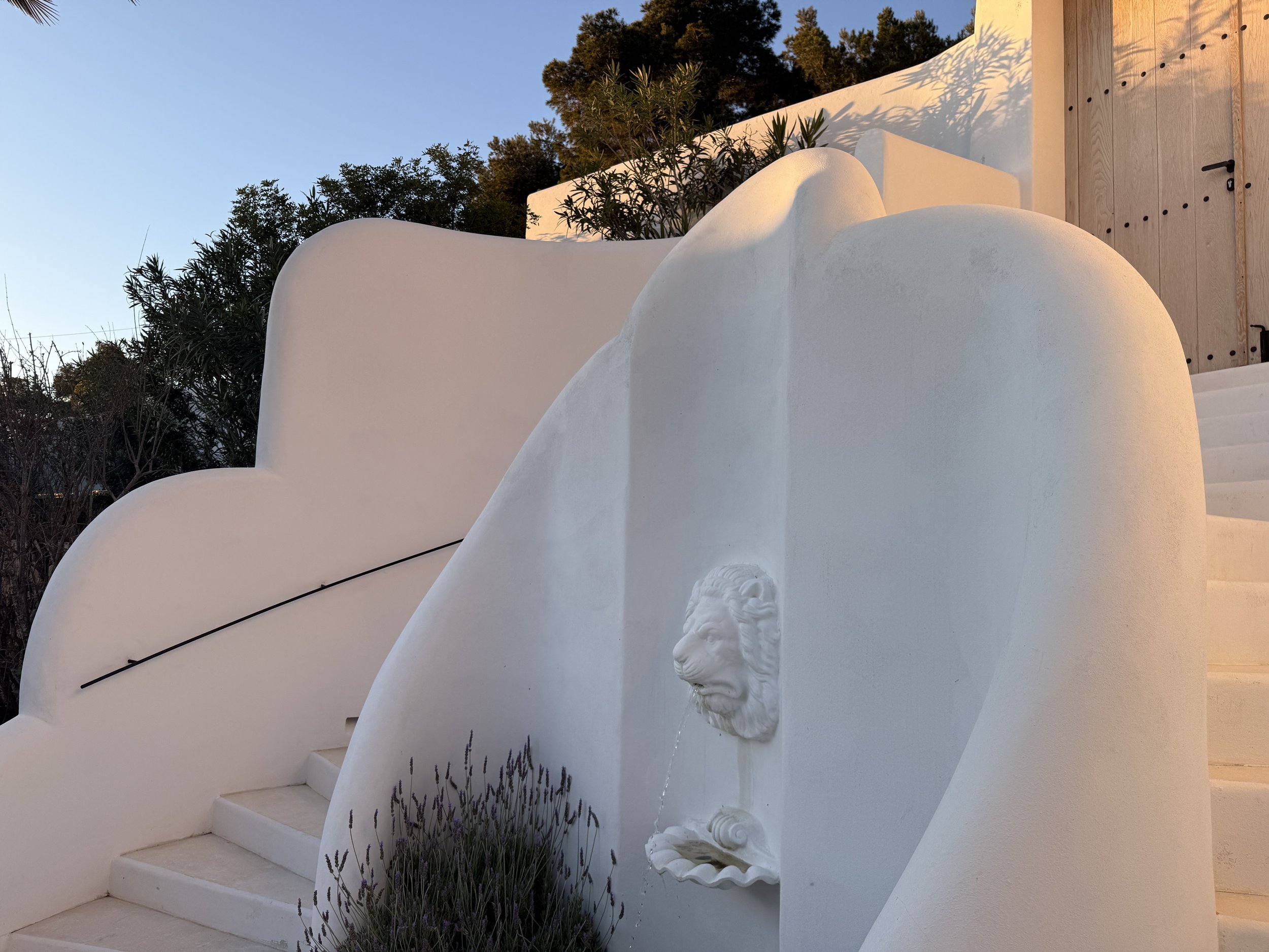 White curved architectural structure with steps, a lion fountain, and outdoor plants against a clear blue sky.