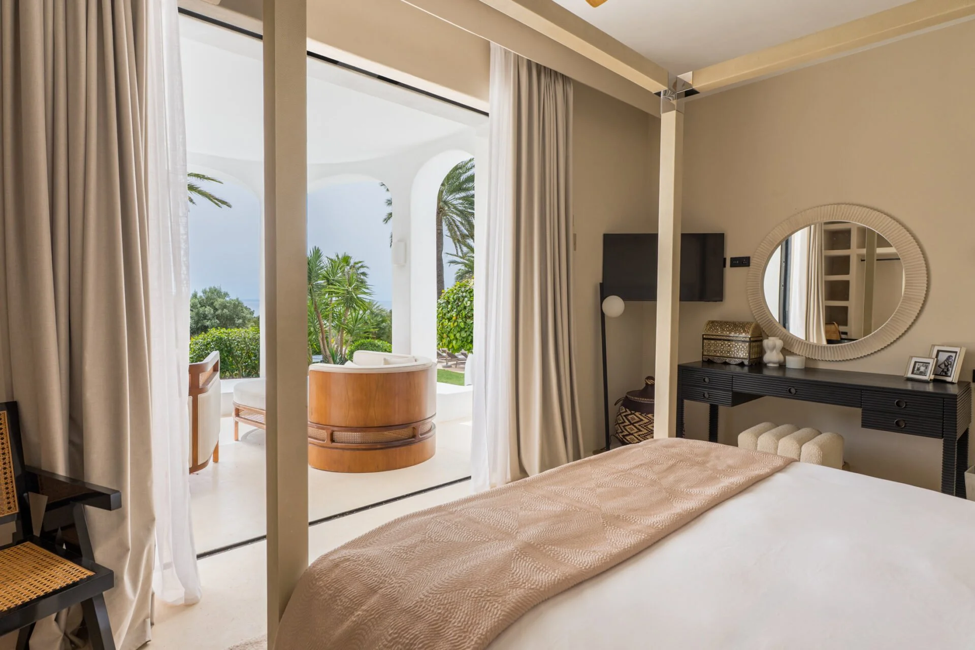 A hotel room with a bed, a black dresser with a mirror, flat-screen TV, and decorative items on top. The room opens to a balcony with a white lounge chair and view of palm trees and a blue sky.