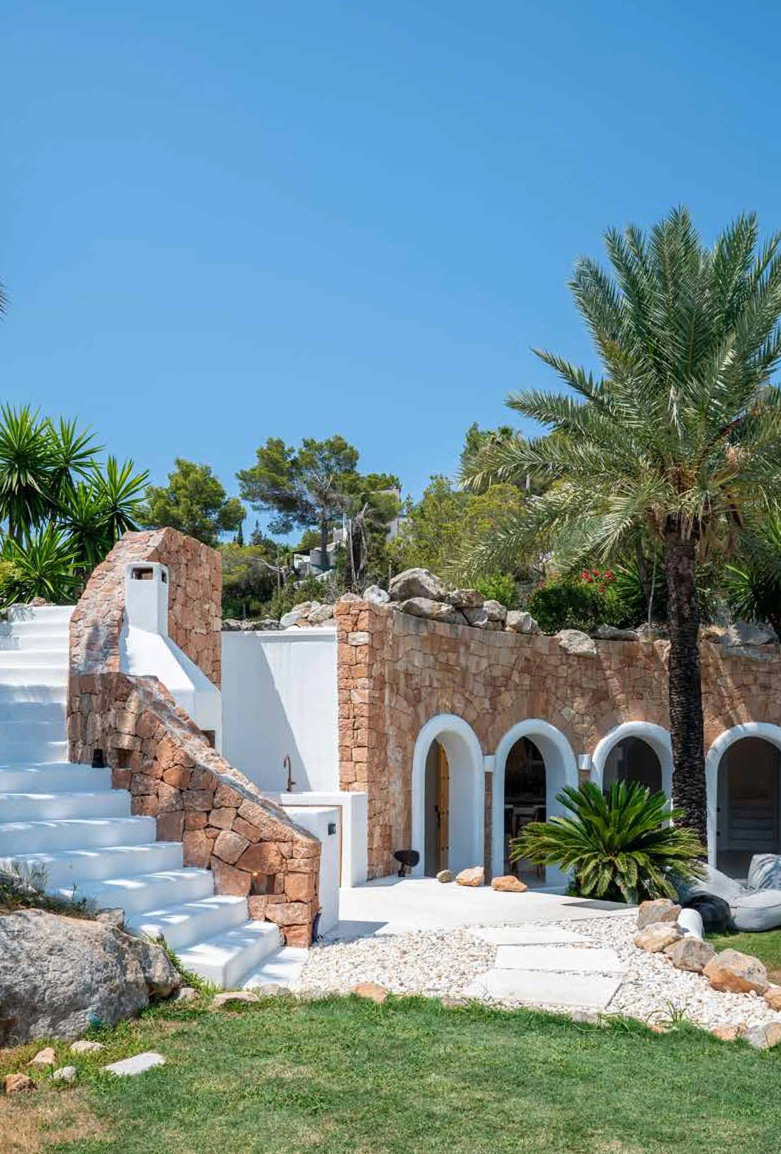 A Mediterranean-style outdoor space with white stairs, a stone wall, arched doorways, palm trees, and lush greenery under a clear blue sky.