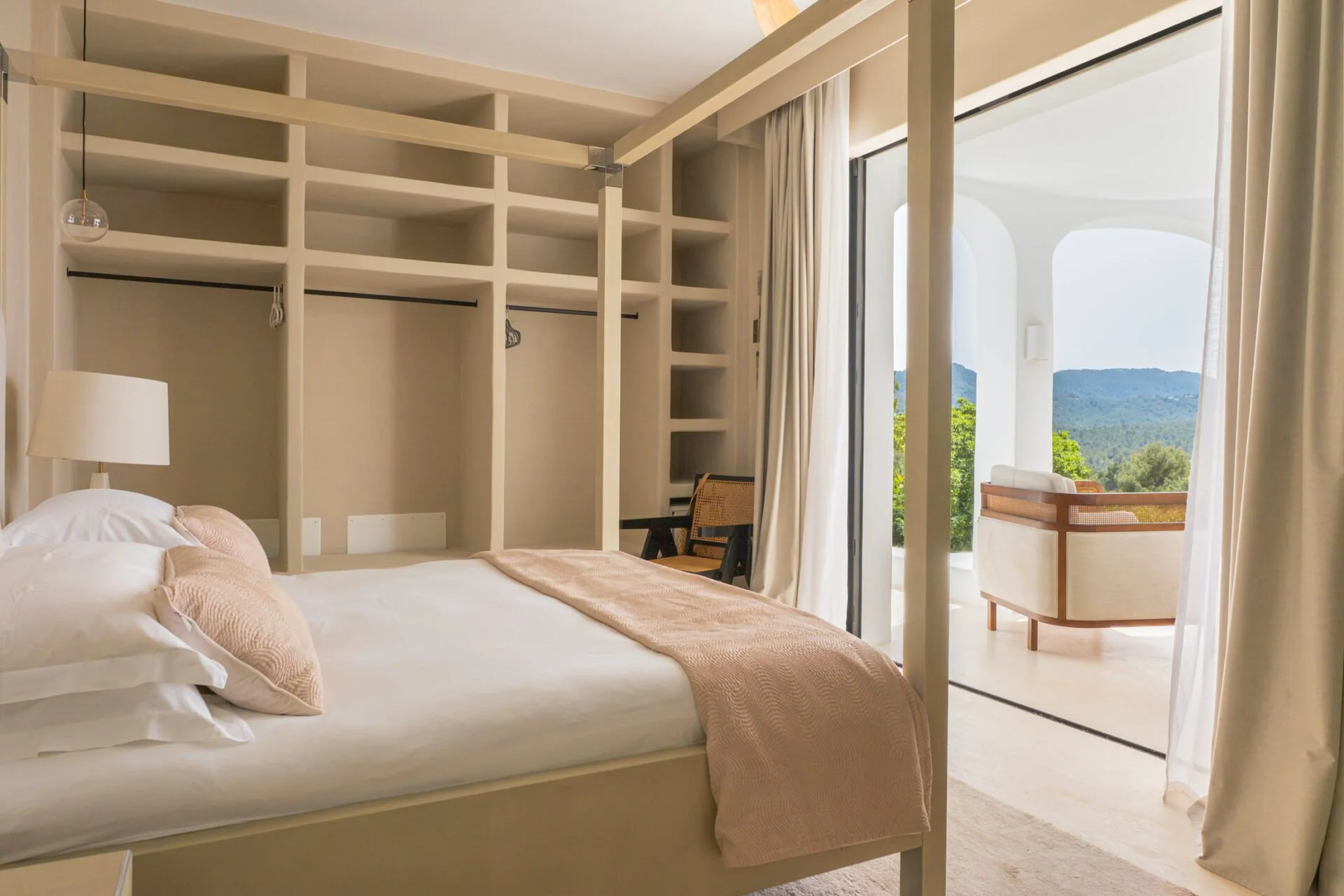 Bedroom with a white bed, beige pillows, and a pink blanket, open balcony doors revealing a scenic mountain view, and built-in shelving behind the bed.