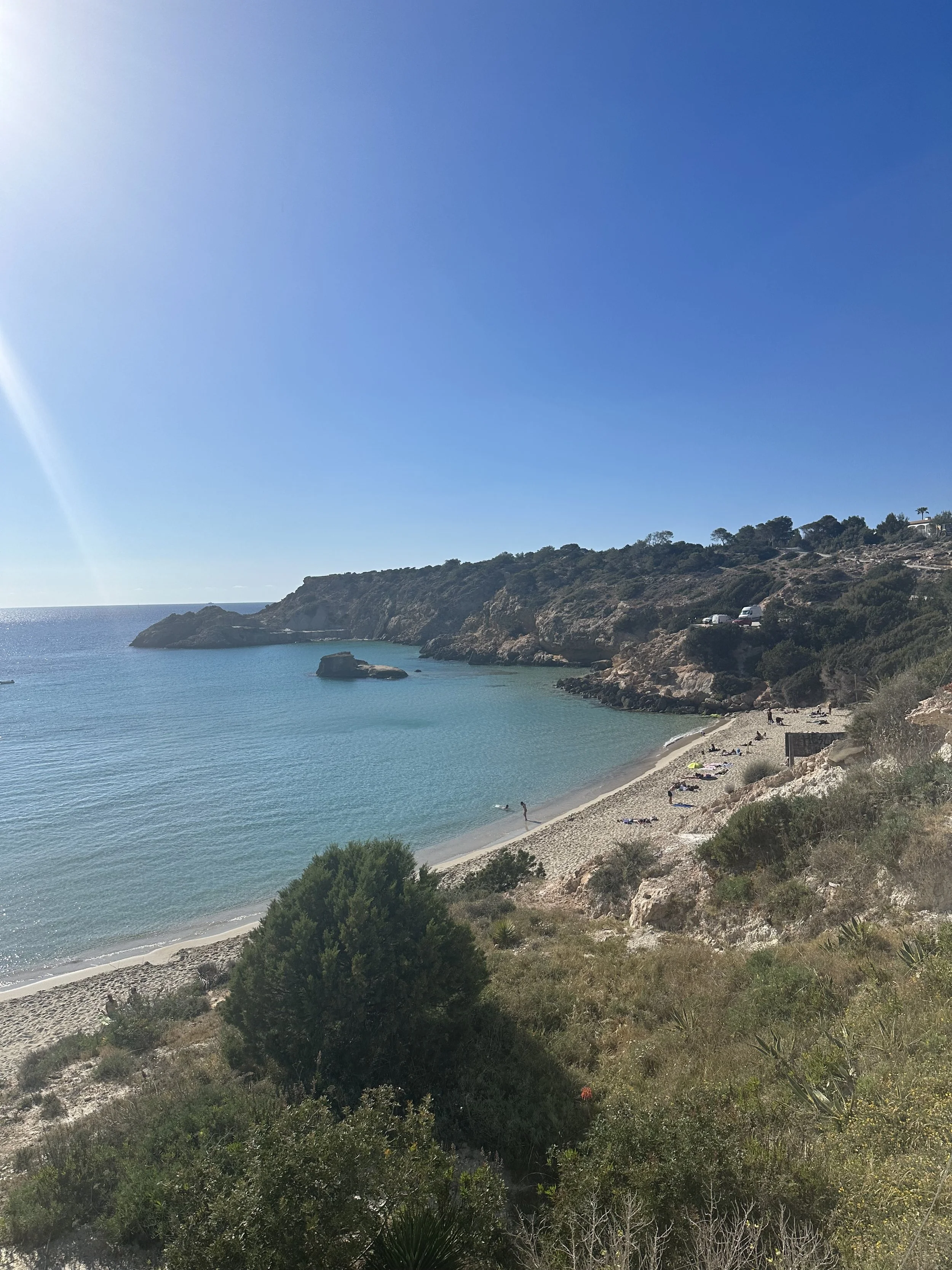Coastal beach with sandy shore, rocky cliffs, and clear blue water under a bright blue sky with minimal clouds.