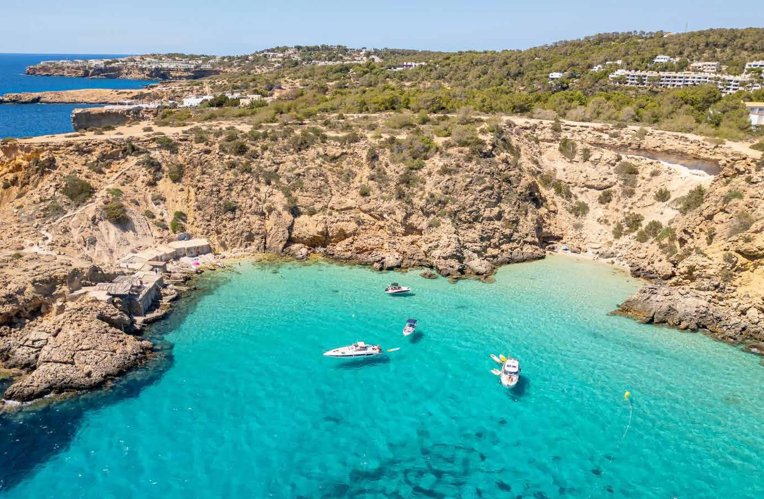 A cove with clear turquoise water surrounded by rocky cliffs and some boats floating in the water. There are a few buildings or huts along the shoreline and a hill with vegetation and houses in the background.