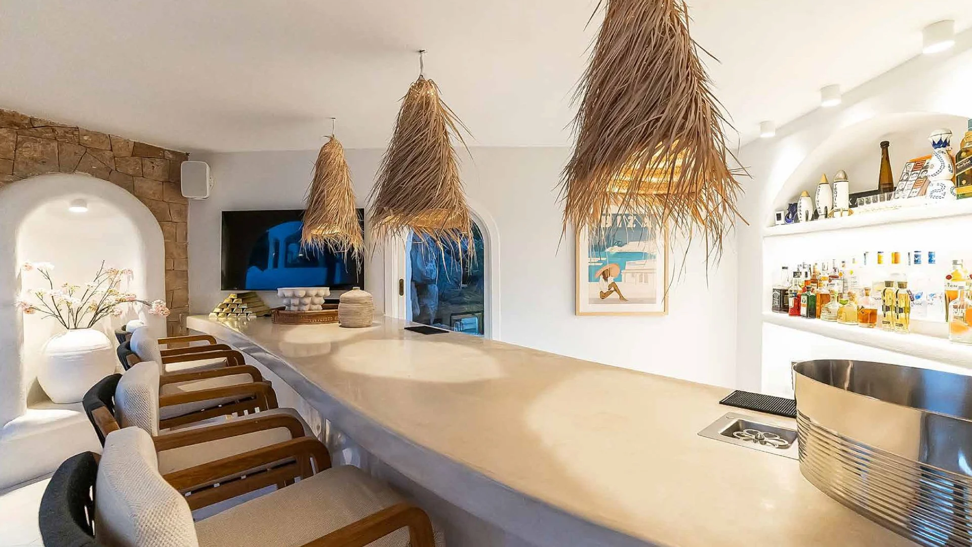 A modern bar with a beige countertop, hanging straw pendant lights, bar stools with beige cushions and wooden arms, a leafy plant in a white vase, shelves with bottles of alcohol, and a framed picture of a person holding an umbrella near a boat on water.