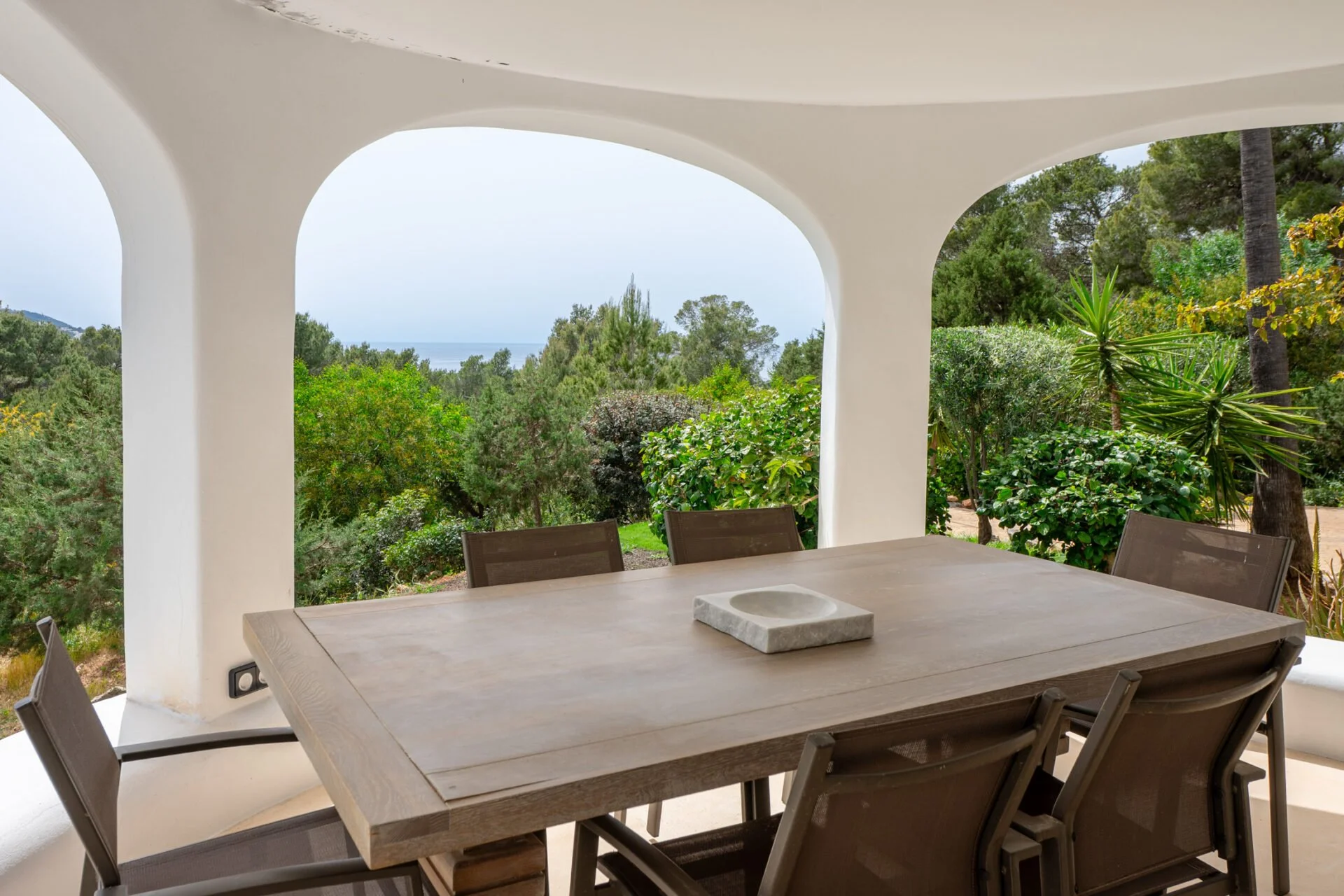 Outdoor patio with a rectangular wooden table and chairs, surrounded by lush green trees and view of the ocean in the distance. White curved architectural columns frame the scene.