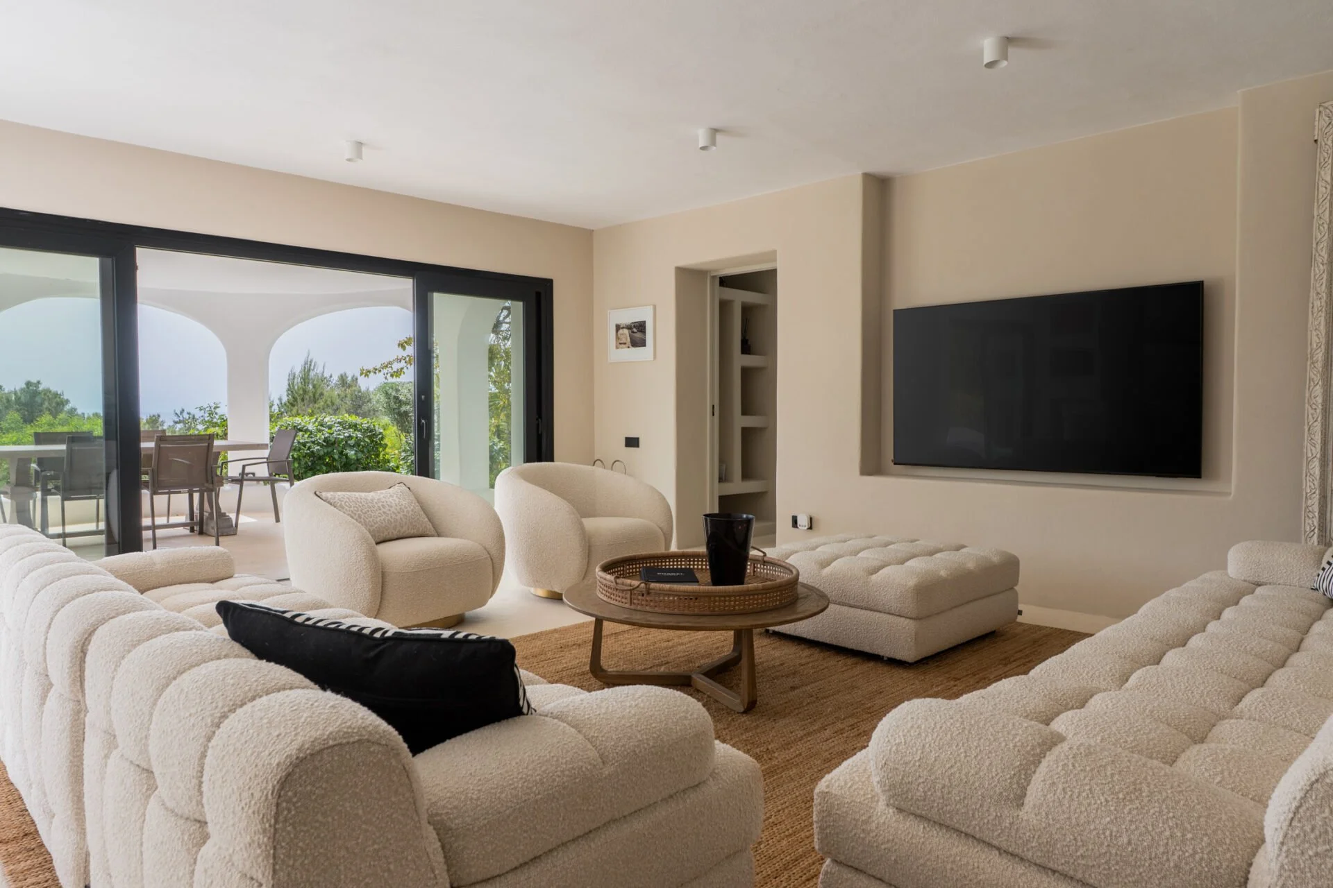 Living room with beige upholstered sectional sofa, two matching armchairs, black pillow, round wooden coffee table with black vase, and wall-mounted flat screen TV, with sliding glass door showing outdoor patio and greenery.