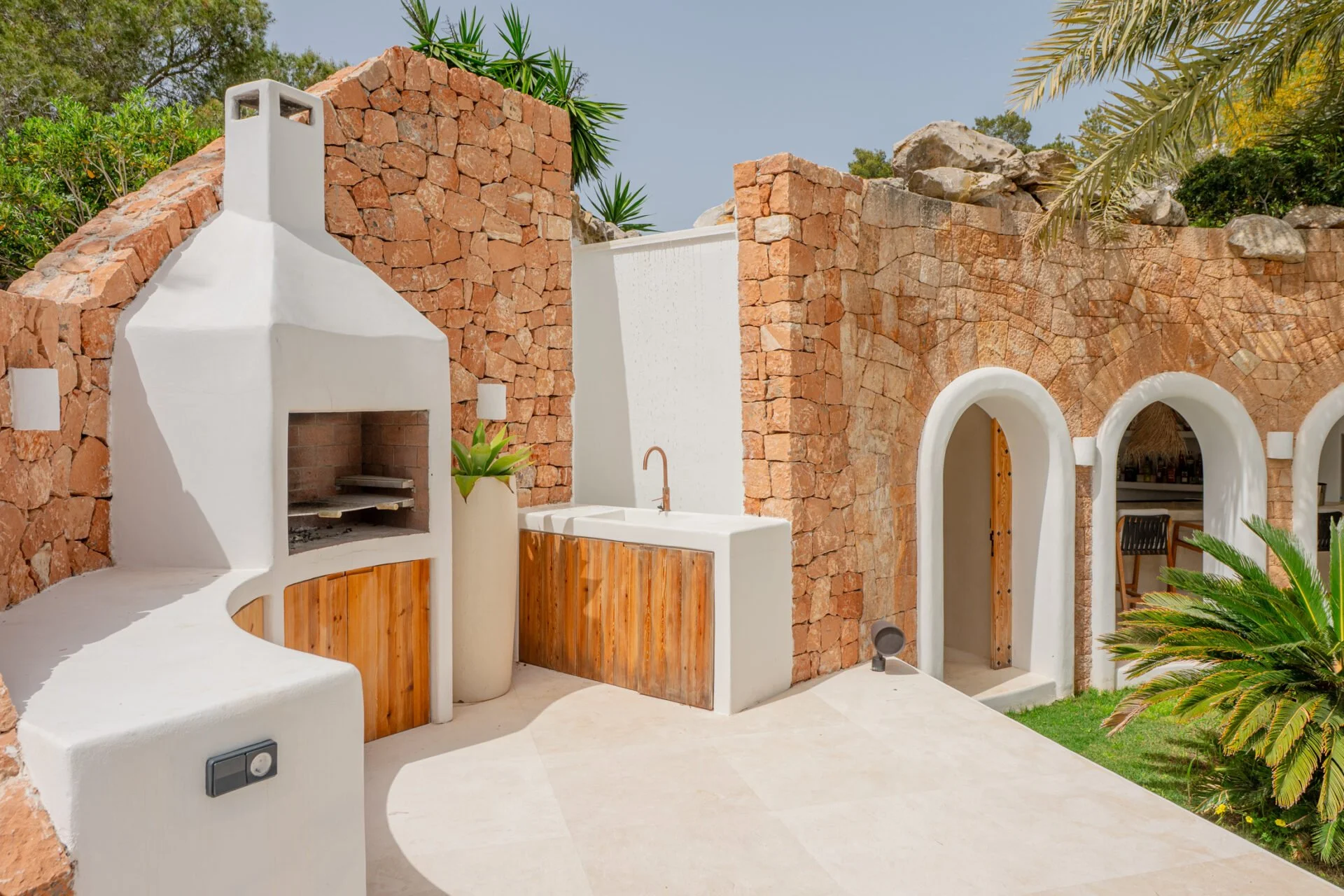 A courtyard with white stucco and brick walls, built-in outdoor fireplace, potted plant, and open archways leading to a dining area; surrounded by lush green plants and trees.