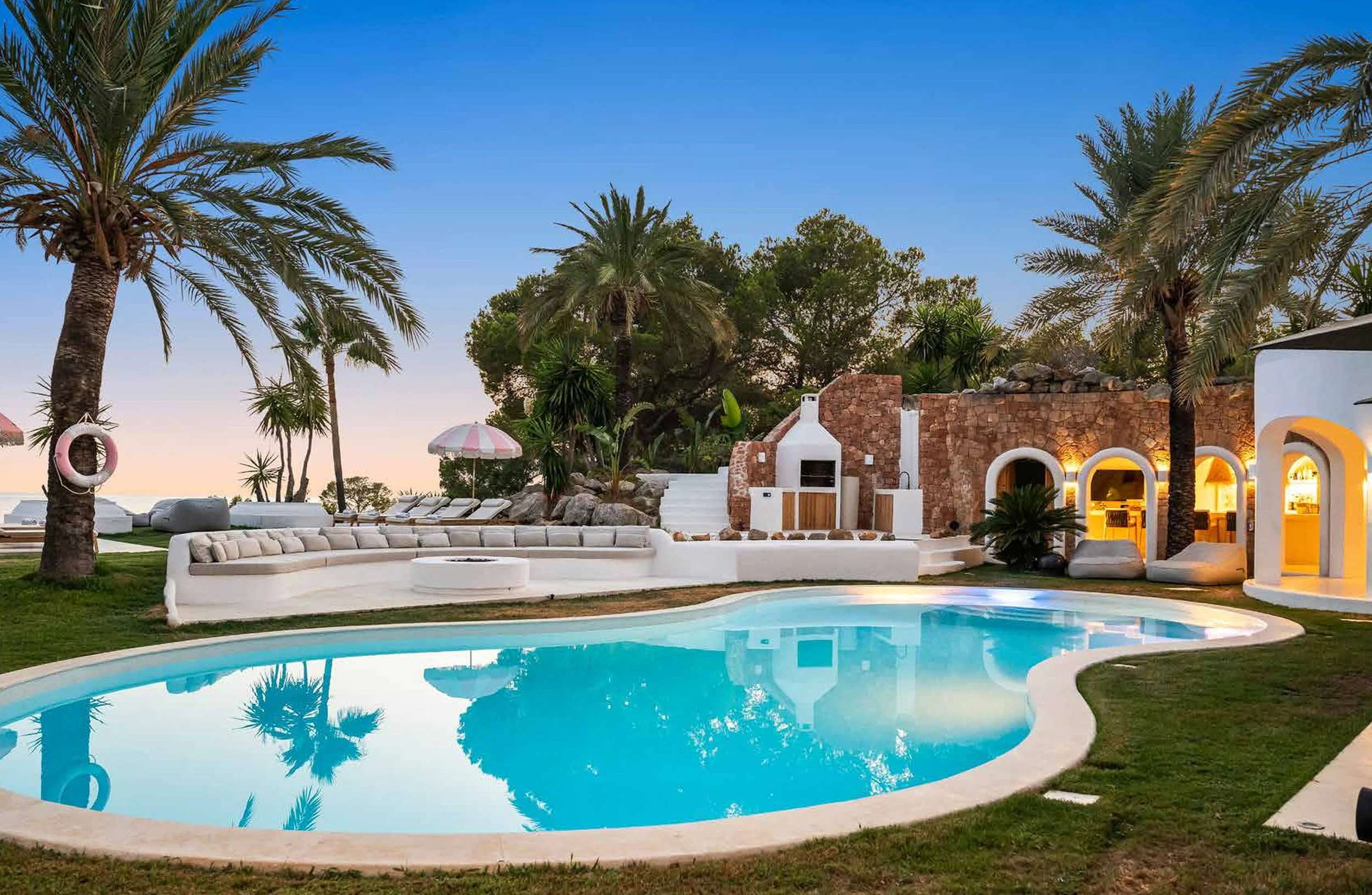 A luxurious backyard with a swimming pool, surrounded by palm trees, white lounge chairs, an outdoor fireplace, and a cozy sitting area with an arched white structure at sunset.
