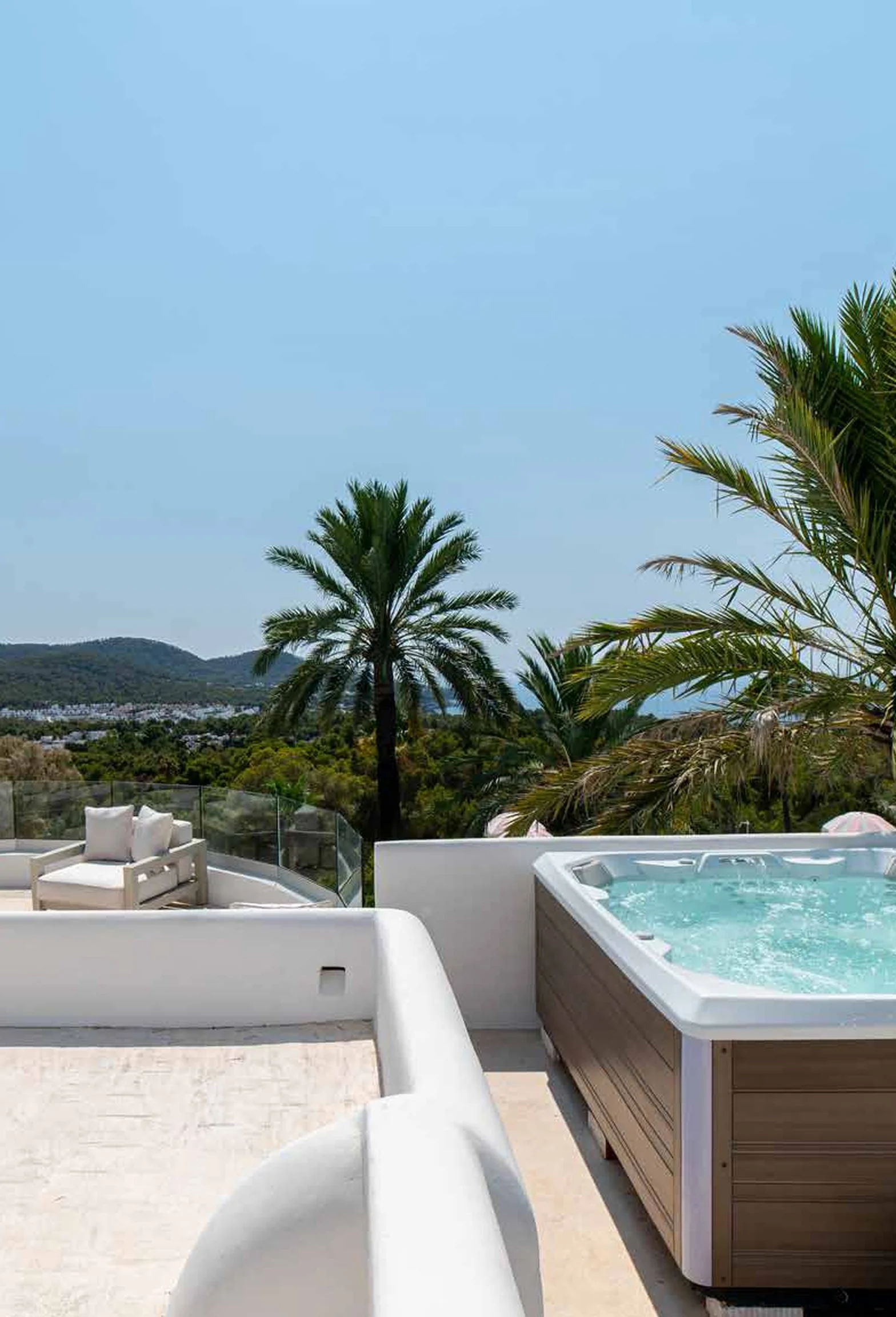 Luxury balcony with hot tub and white outdoor furniture, overlooking hills and palm trees under a clear blue sky.