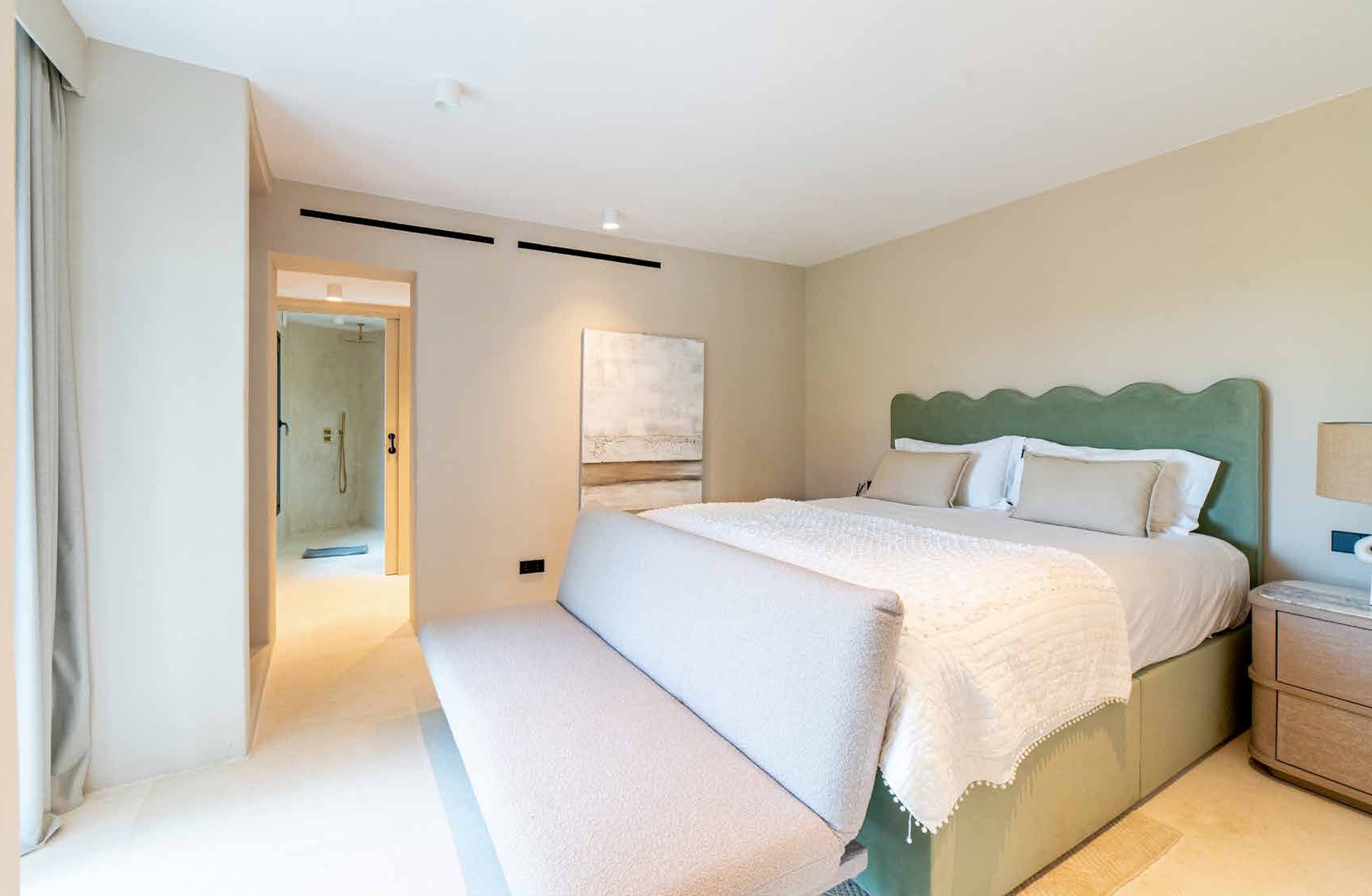 Hotel room with a large bed, green headboard, white bedding, and beige walls. A bench is at the foot of the bed, and a nightstand with a lamp is on the right side. An open doorway leads to a bathroom with a shower.