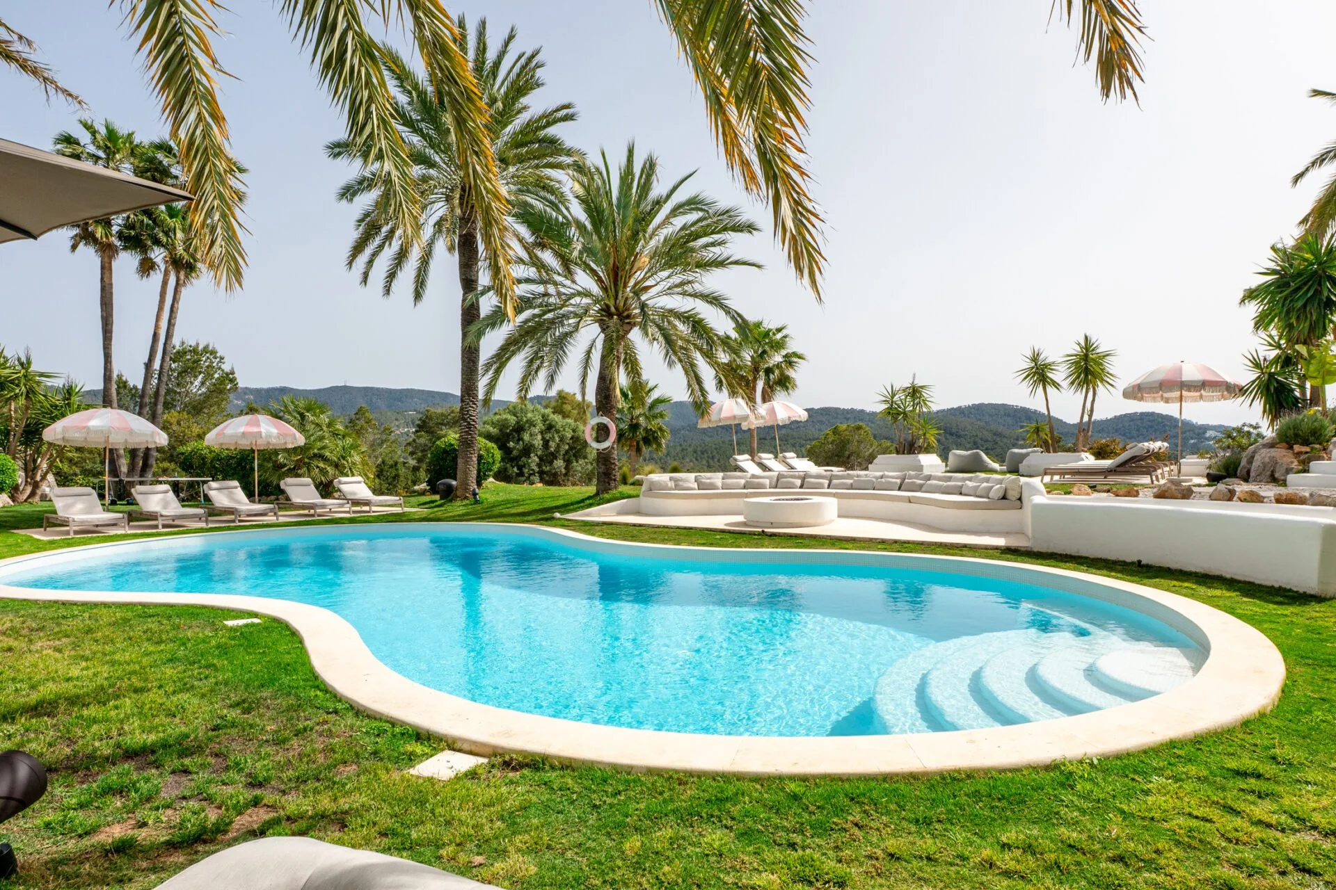 Poolside area with lounge chairs, parasols, and palm trees in a sunny outdoor setting overlooking mountains.