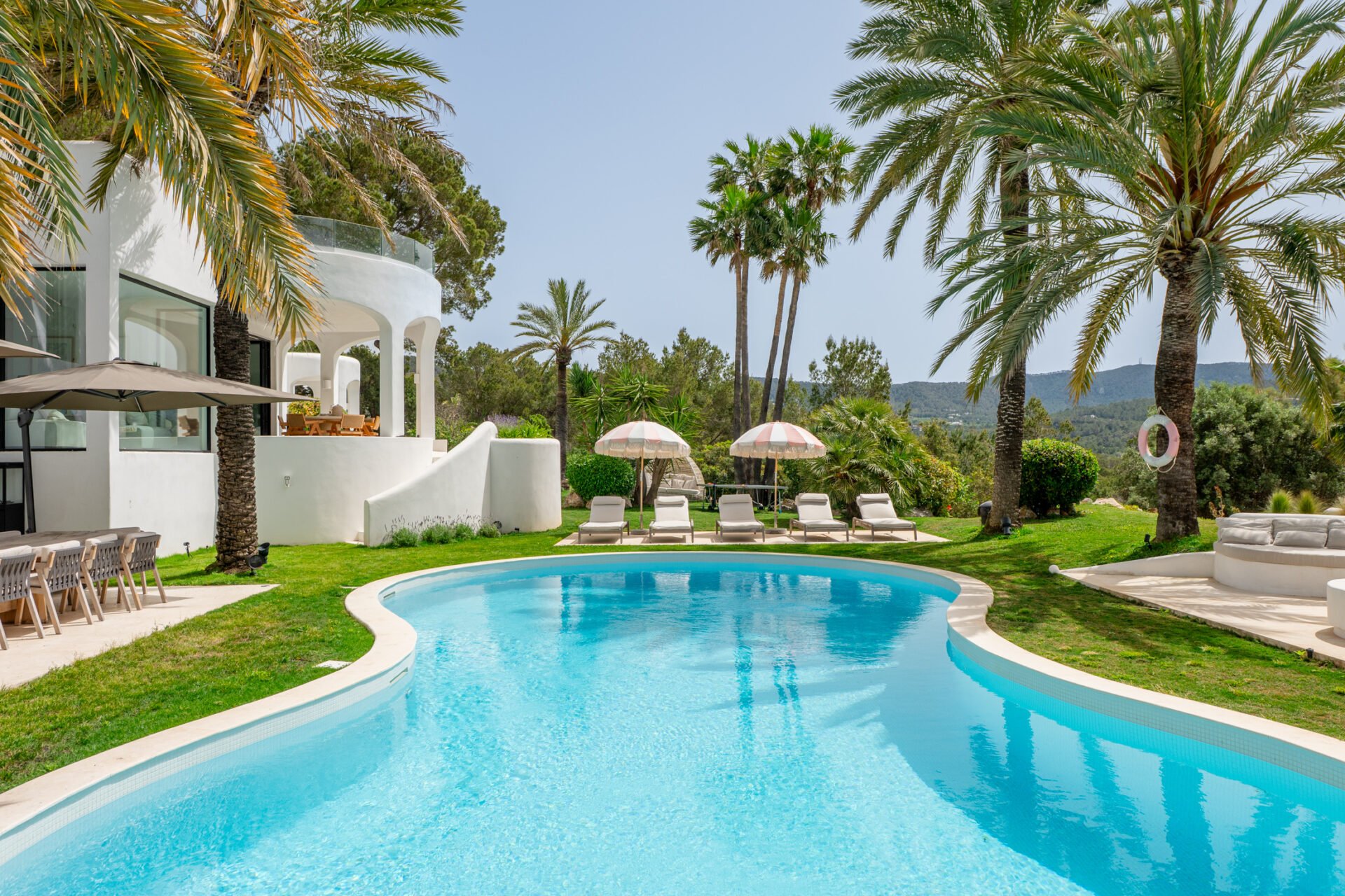 A luxurious outdoor pool in Ibiza area surrounded by palm trees, lounge chairs with umbrellas, and a modern white building with curved architecture, set against a backdrop of green hills and a clear blue sky.