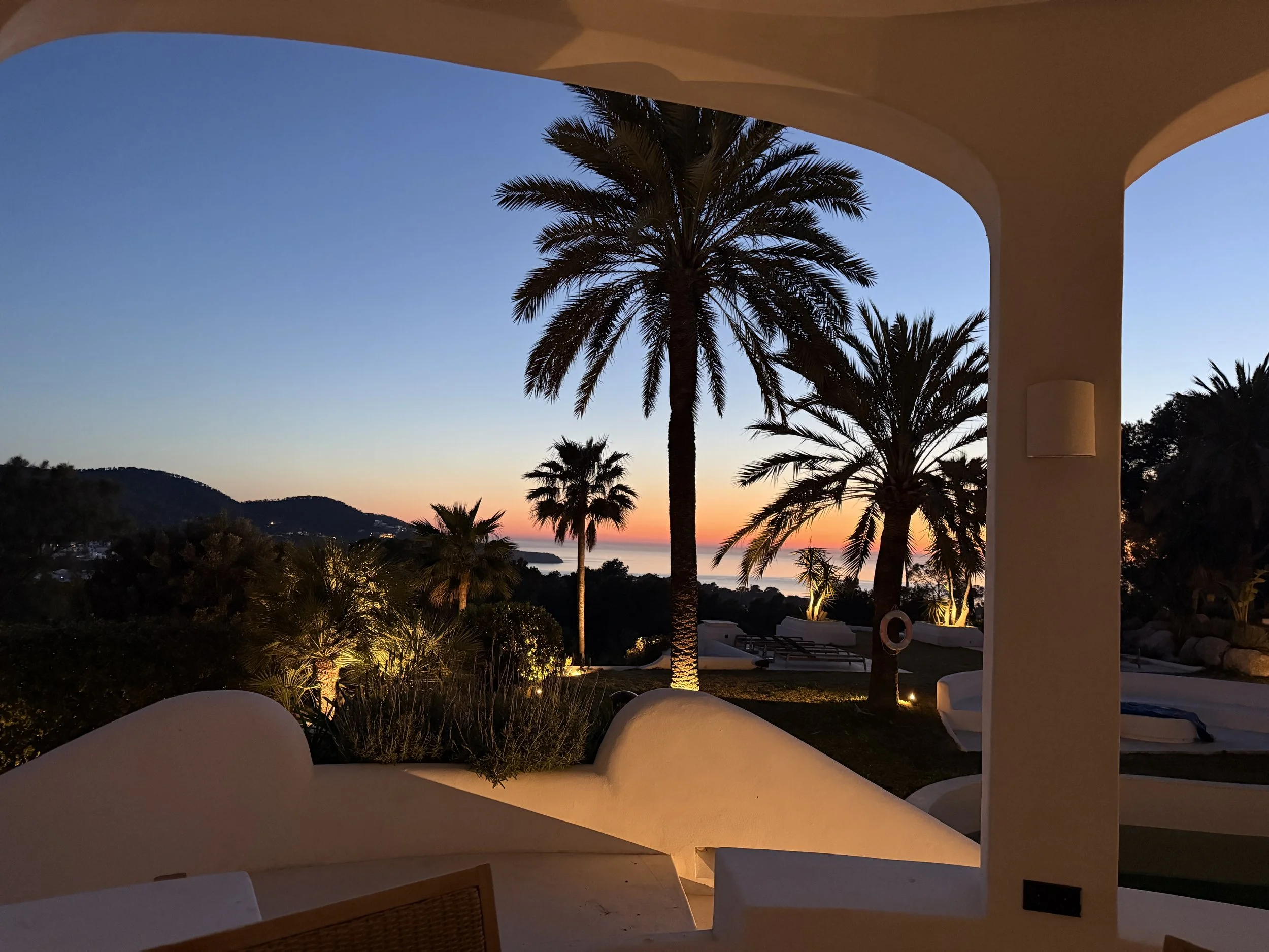 Sunset view with palm trees over the ocean seen from a terrace with white furniture and architecture.