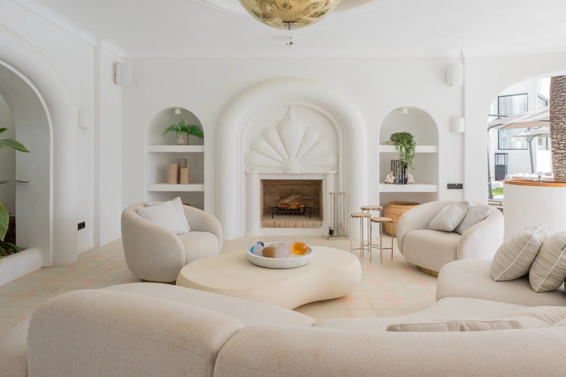 A cozy living room with a white color scheme, featuring a white curved sofa, a circular white coffee table with decorative items, a white fireplace with built-in shelves, and minimalist decor including potted plants and books.