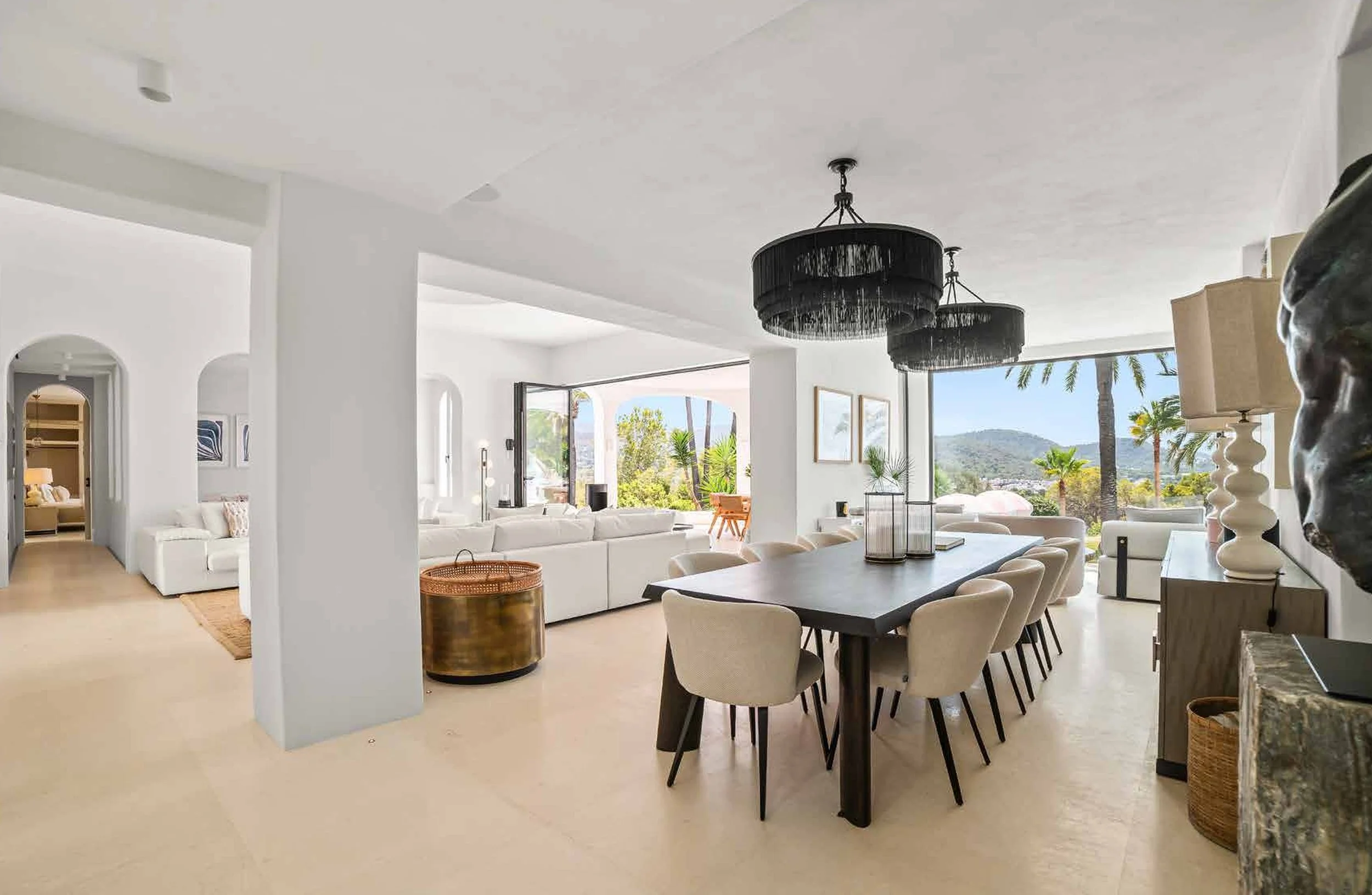 Wide view of a bright, modern living and dining area with large windows showcasing a scenic outdoor landscape, white walls, and neutral-colored furniture, including a large dining table with chairs and a spacious white sectional sofa.