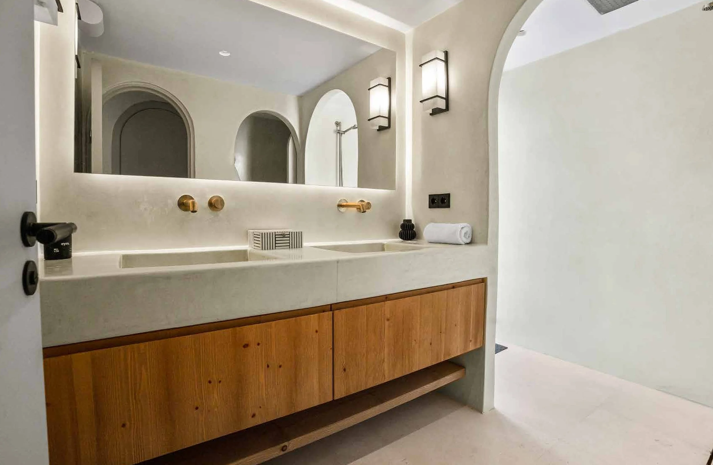Modern bathroom with a large mirror, double vanity sink with wooden cabinet, gold fixtures, and minimalist decor including a rolled towel, a small black vase, and black wall lights.