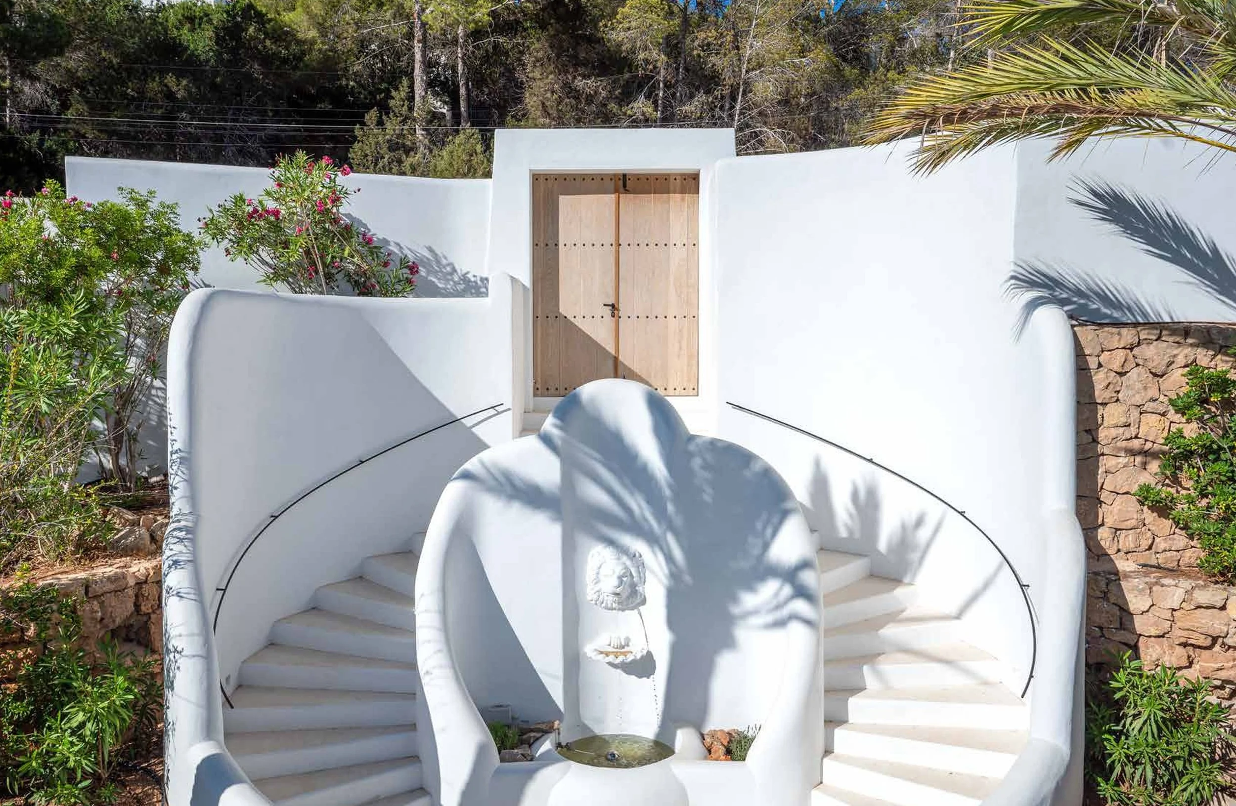 White outdoor staircase and fountain with palm tree shadows, surrounded by plants and a stone and white wall backdrop.