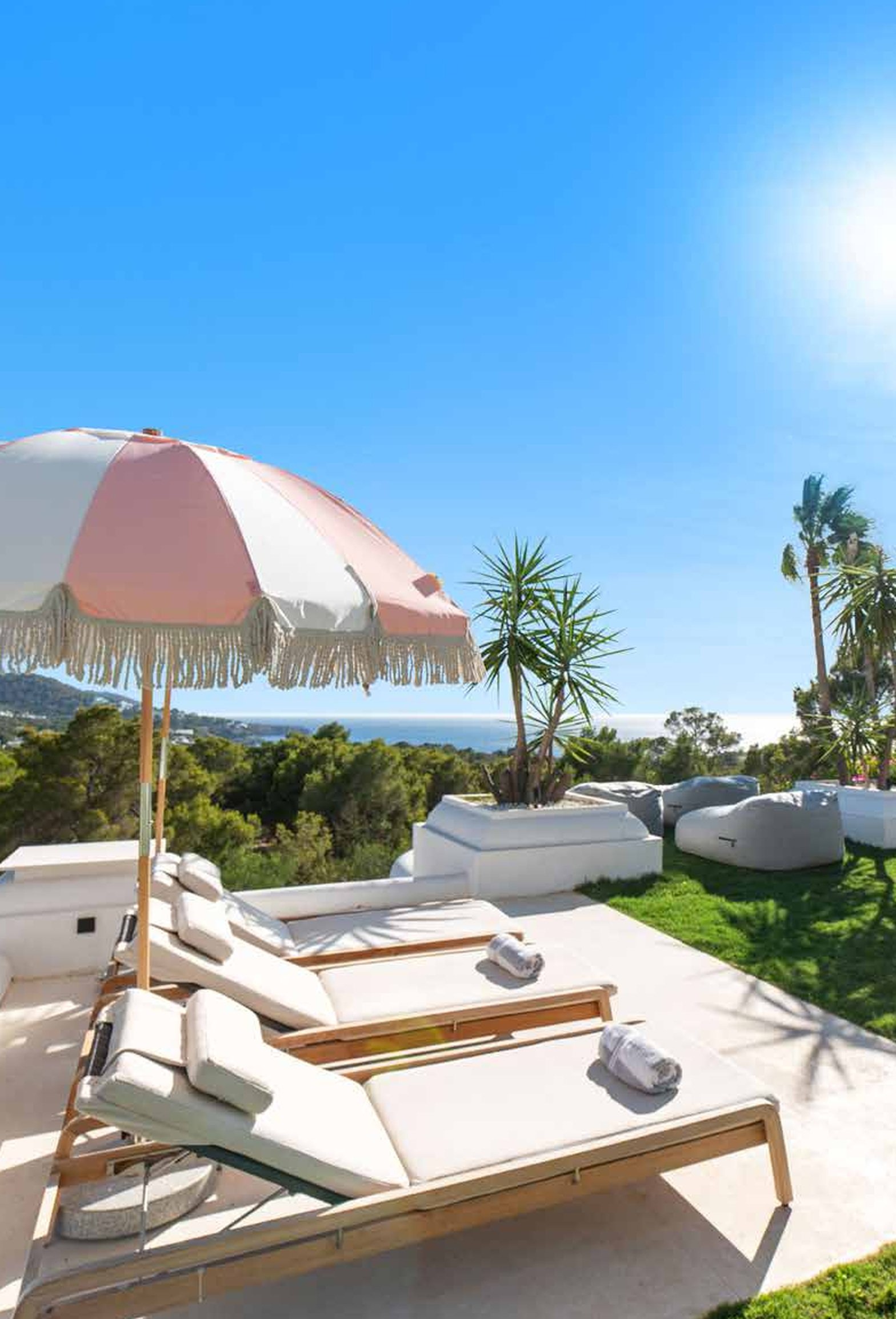 A poolside lounge area with two beige lounge chairs, rolled towels, a pink and white beach umbrella, and tropical plants under a clear blue sky with the sun shining.