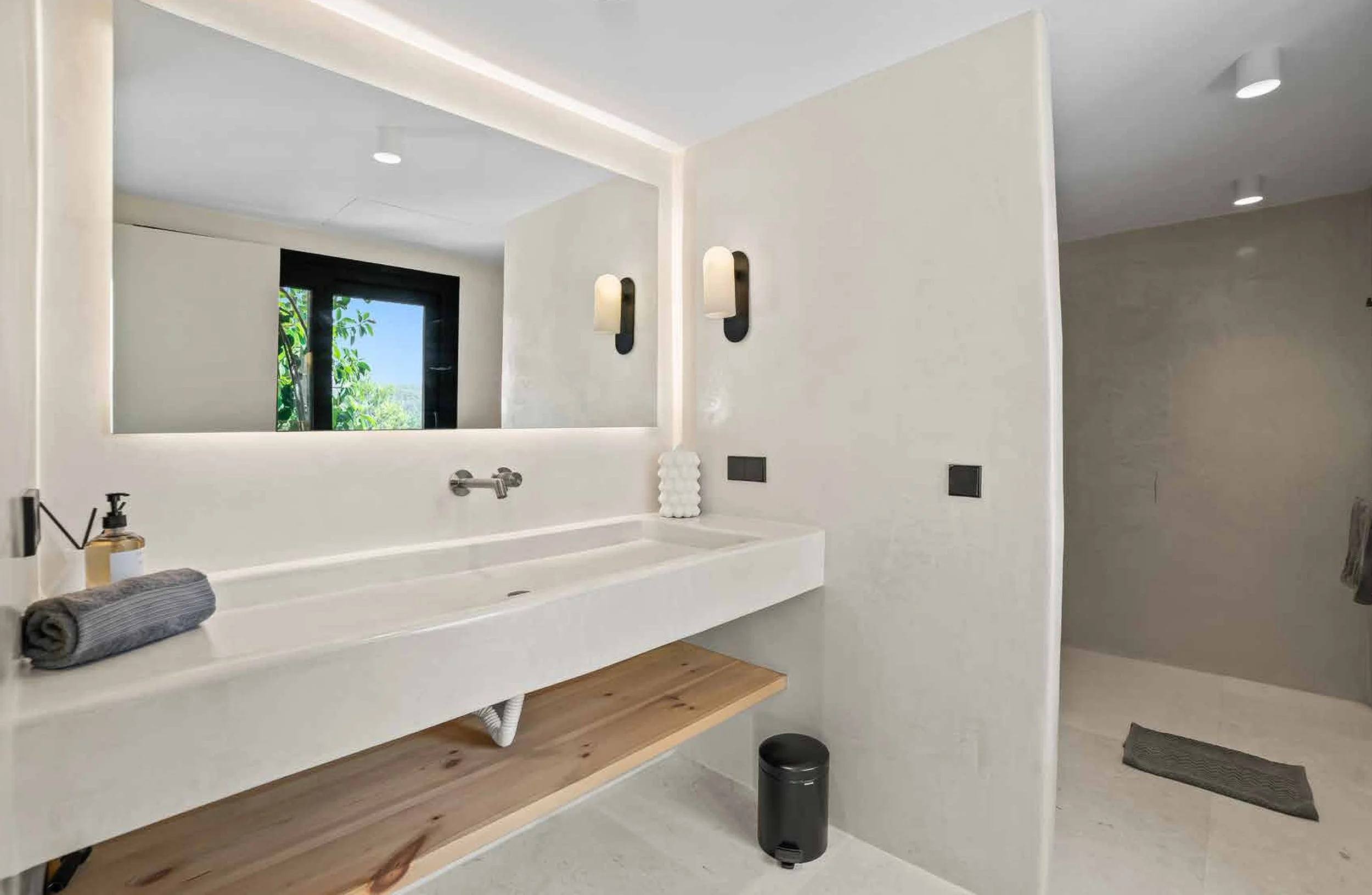 Modern bathroom with a large white sink, wooden shelf, grey towel, soap dispenser, mirror, black wall lights, and a shower area with grey wall and floor tiles.