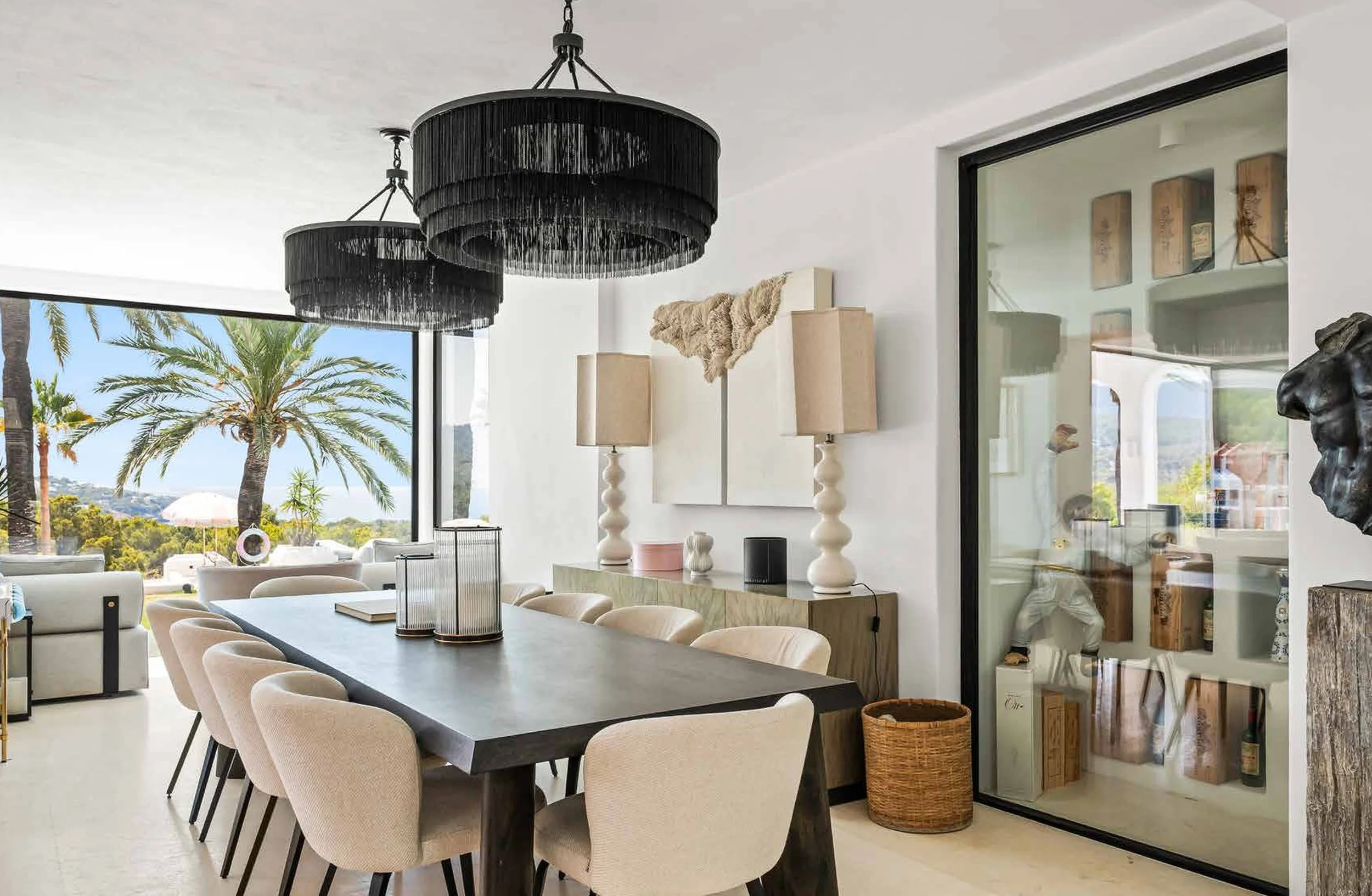 Modern dining room with large black chandelier, beige chairs around a dark wooden table, tropical plants outside, and decorative lamps and art inside.
