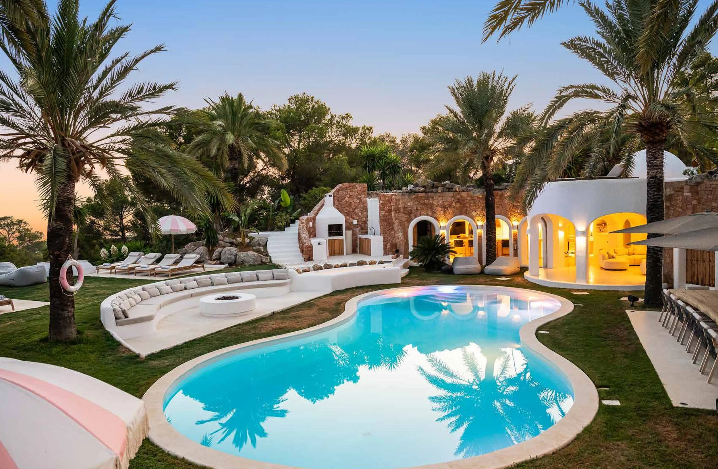 Luxury backyard with a kidney-shaped swimming pool surrounded by palm trees, lounge chairs, and outdoor seating areas at sunset with warm lighting.