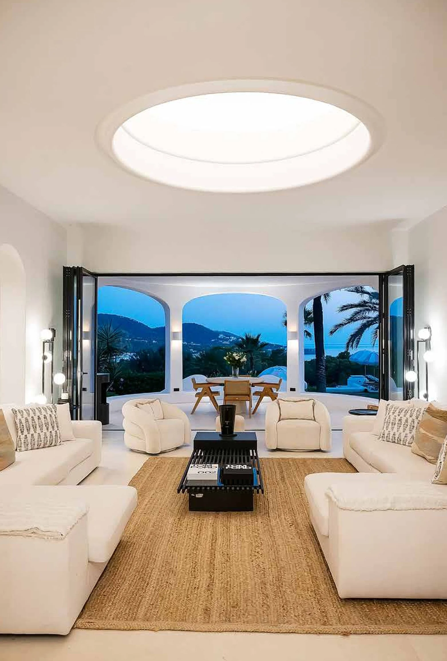 A living room with white sofas, a black coffee table, and a rug, opening to an outdoor patio with a dining table and chairs, overlooking a tropical landscape with palm trees and mountains at dusk.