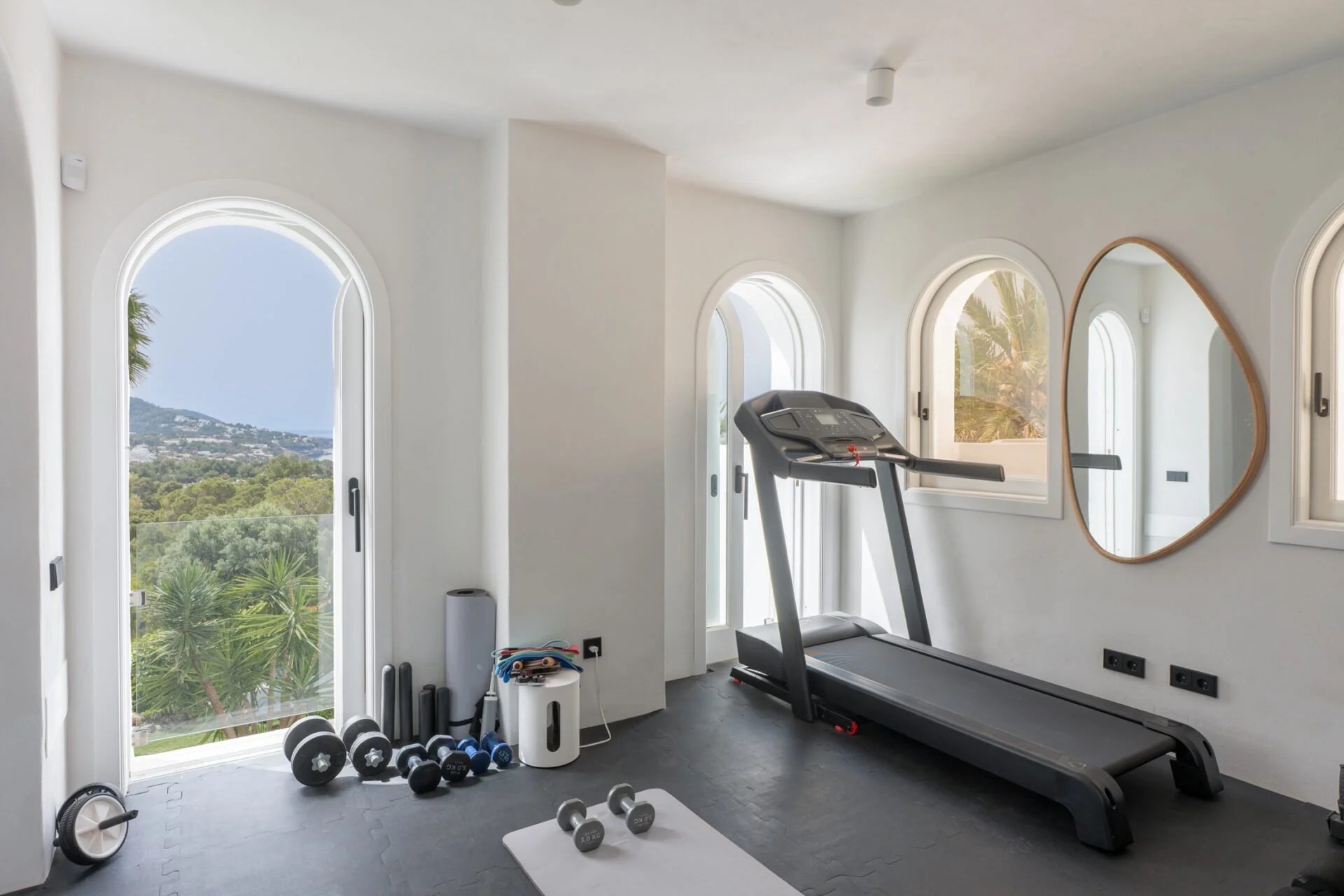 Bright home gym with black treadmill near white wall with arched windows, offering a view of trees and a hillside, with dumbbells, yoga mats, and other workout equipment on dark flooring.