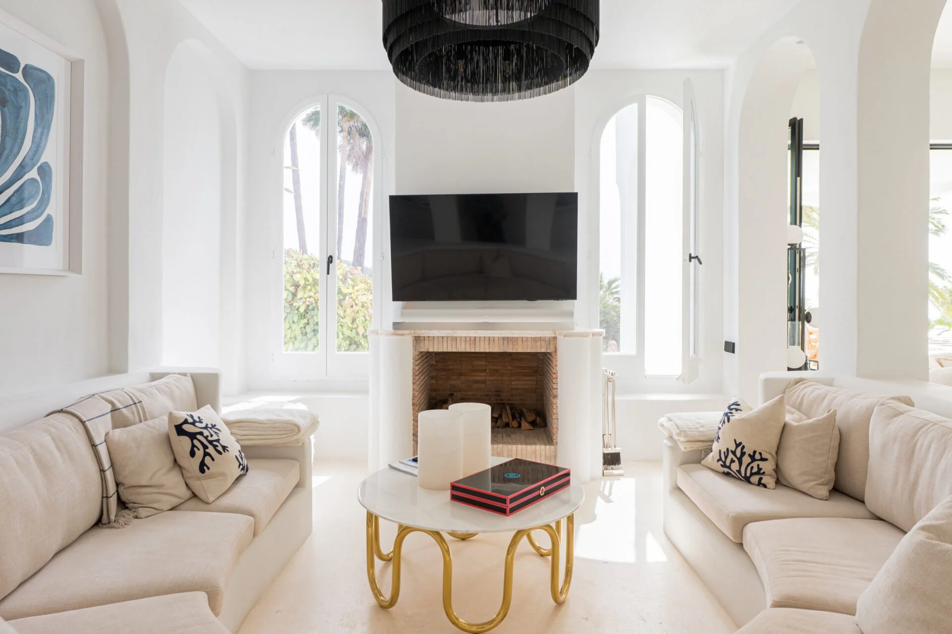 Bright living room with white walls and large arched windows. Two cream-colored sofas with decorative pillows are facing each other, with a round white coffee table in between. The coffee table holds two white candles and a black-and-red box. A black