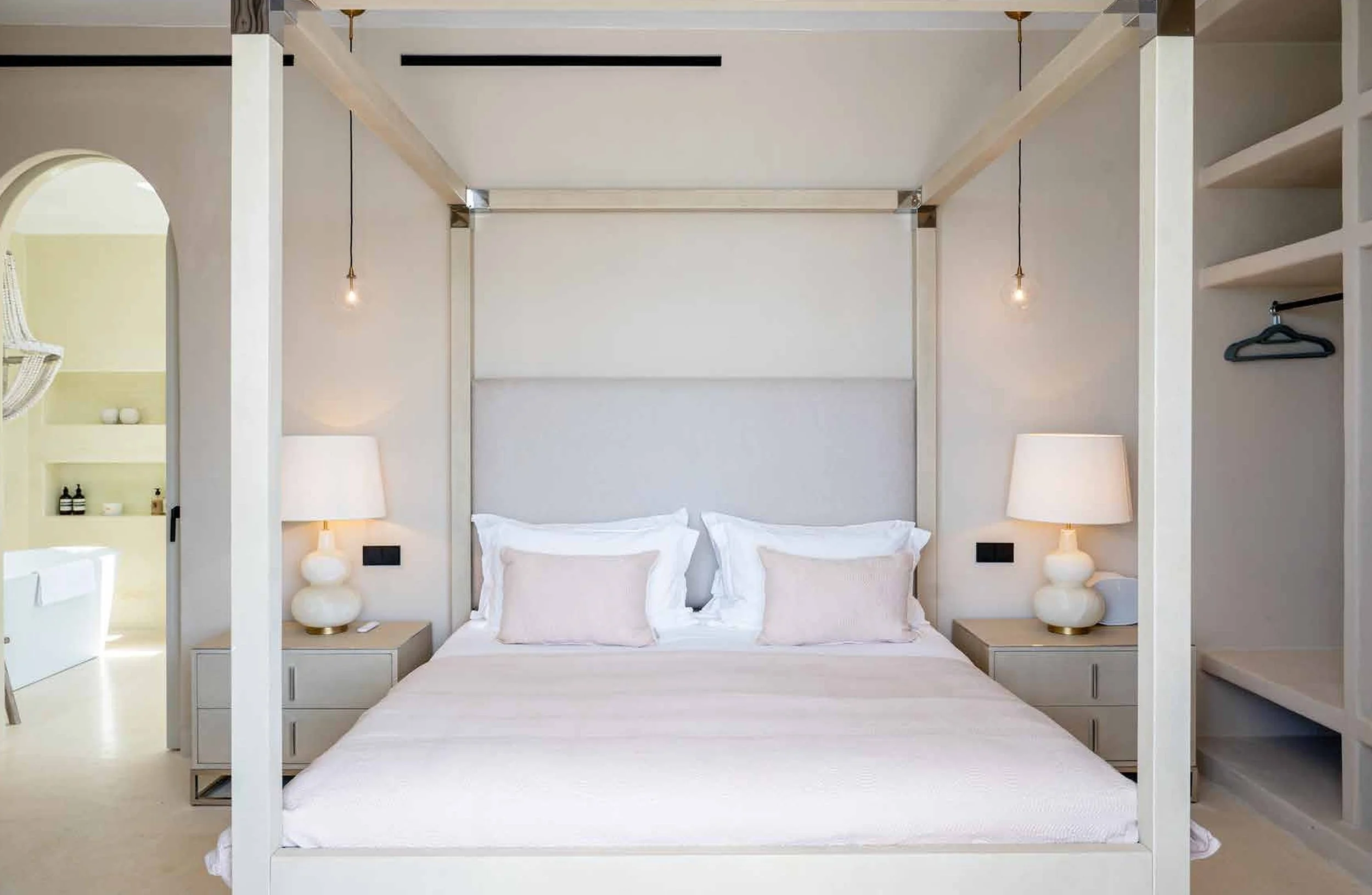 A modern, minimalist bedroom with a large bed, white linens, four pink pillows, and two nightstands with lamps. An open closet is on the right, and a doorway leads to a bathroom on the left.