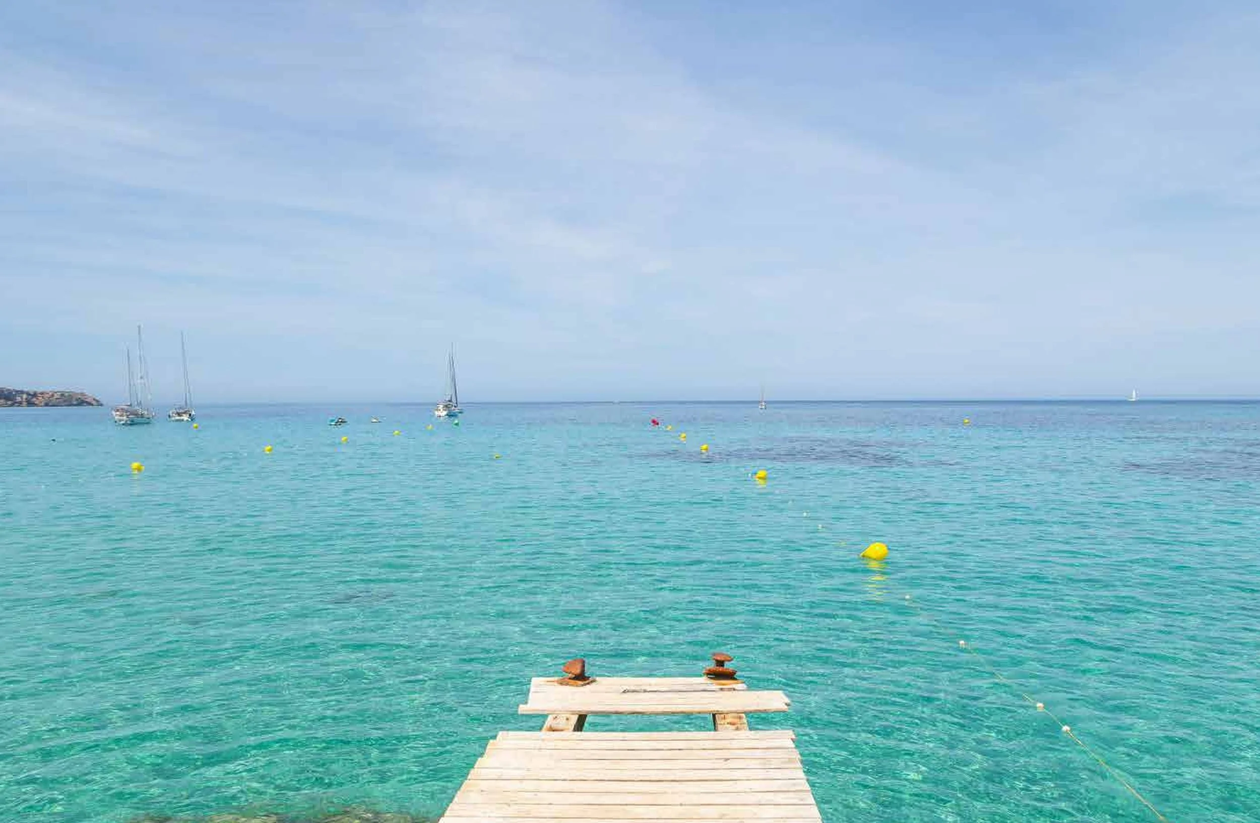 Wooden dock extending into clear turquoise water with sailboats and yellow buoys in the distance under a partly cloudy sky.