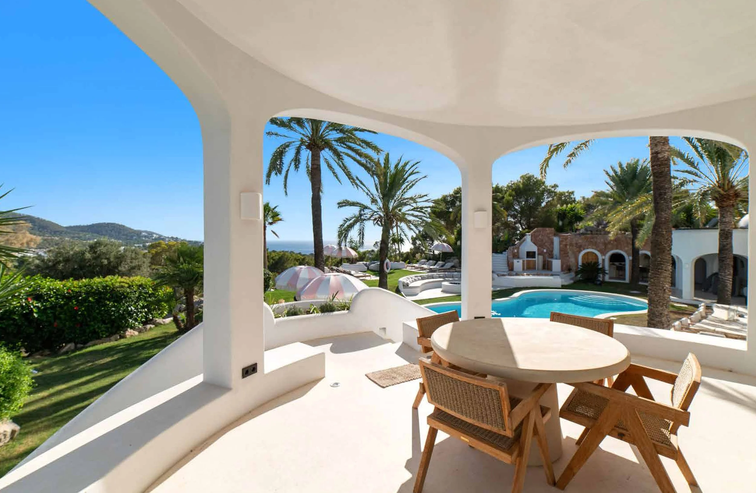 View from a white patio with outdoor dining table, overlooking a pool area with palm trees, umbrellas, and sun loungers in a tropical garden.