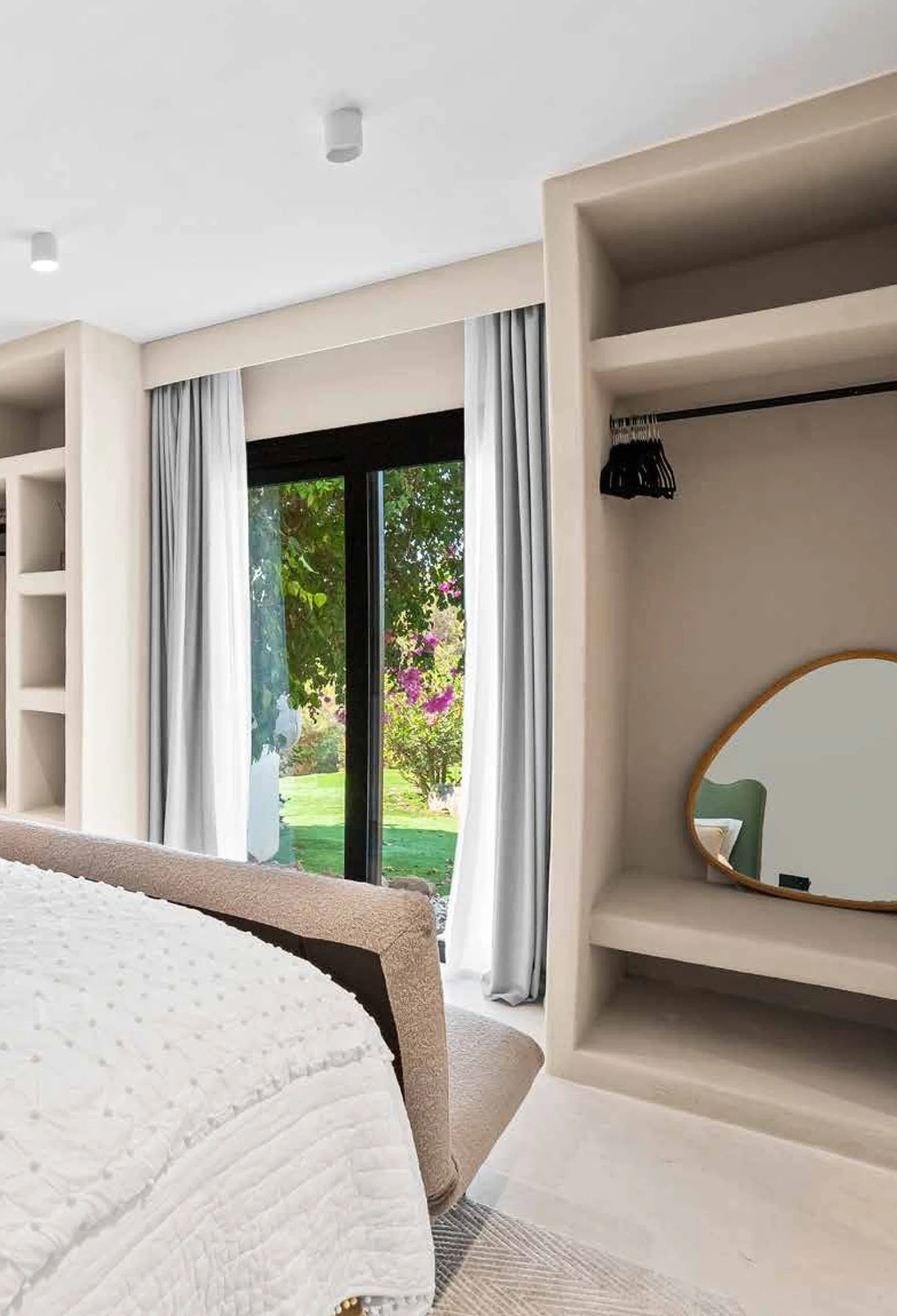 A bedroom with large sliding glass doors leading to a garden, white curtains, and a built-in shelf with a mirror and some hangers.