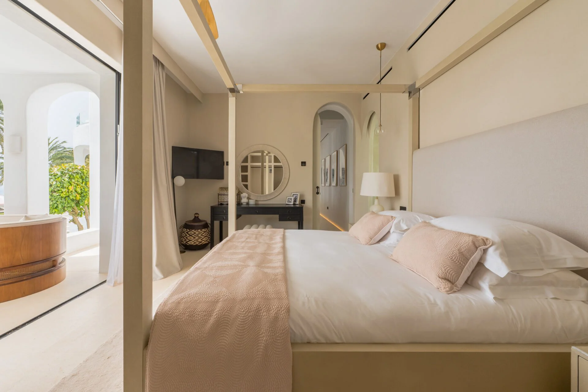 Modern bedroom with a large bed, beige bedding, and pillows; a nightstand with a lamp; a wall-mounted TV; decorative mirror and artwork; patio door with curtains leading outside to greenery.