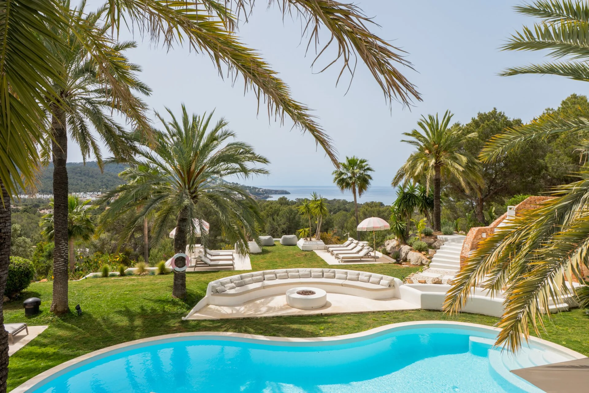 A luxurious outdoor swimming pool surrounded by a lush green lawn and tall palm trees, with lounge chairs and umbrellas overlooking a scenic view of the ocean and distant hills under a clear sky.